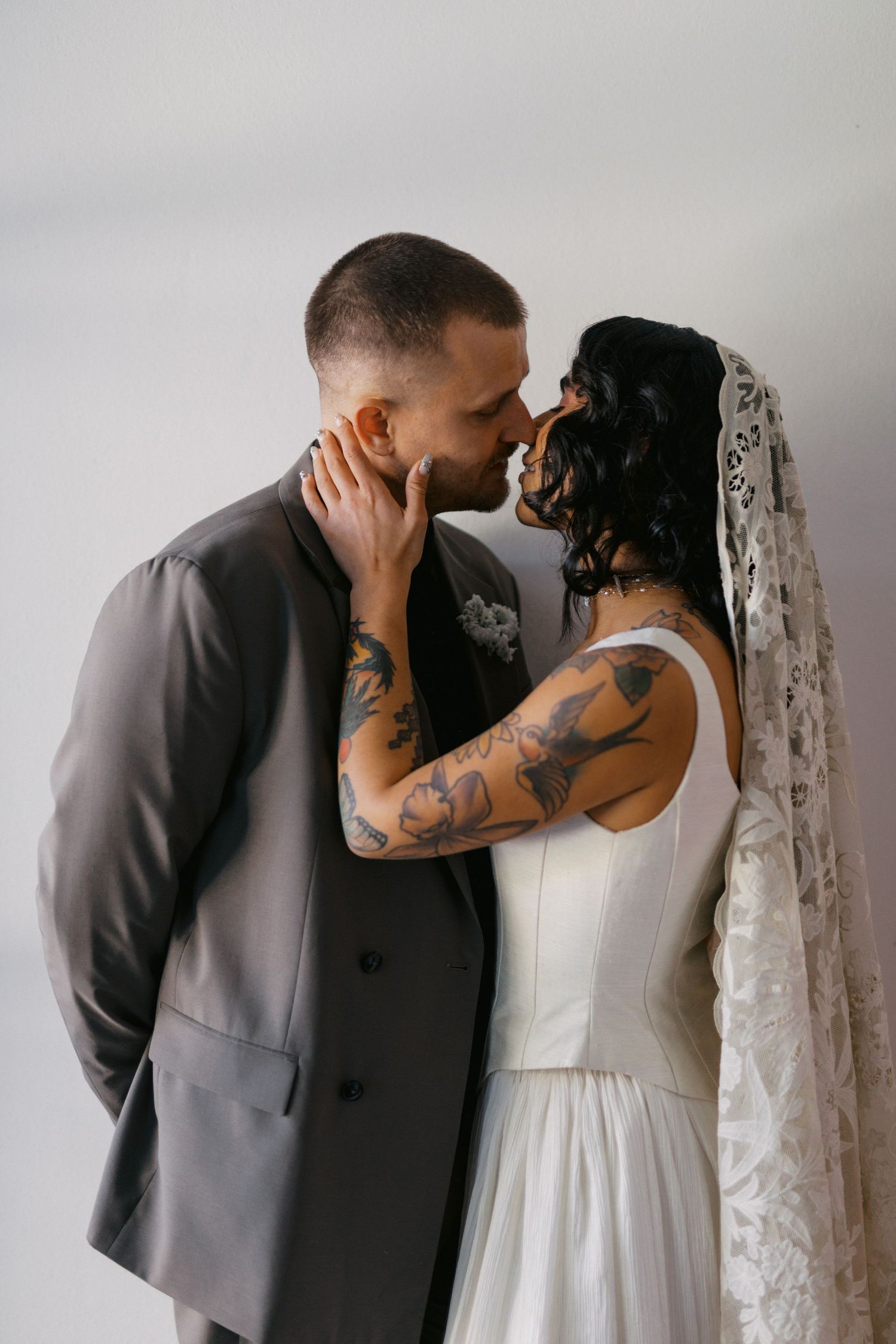Tattooed bride and groom sharing a romantic kiss in wedding attire, bride wearing lace veil and white gown.