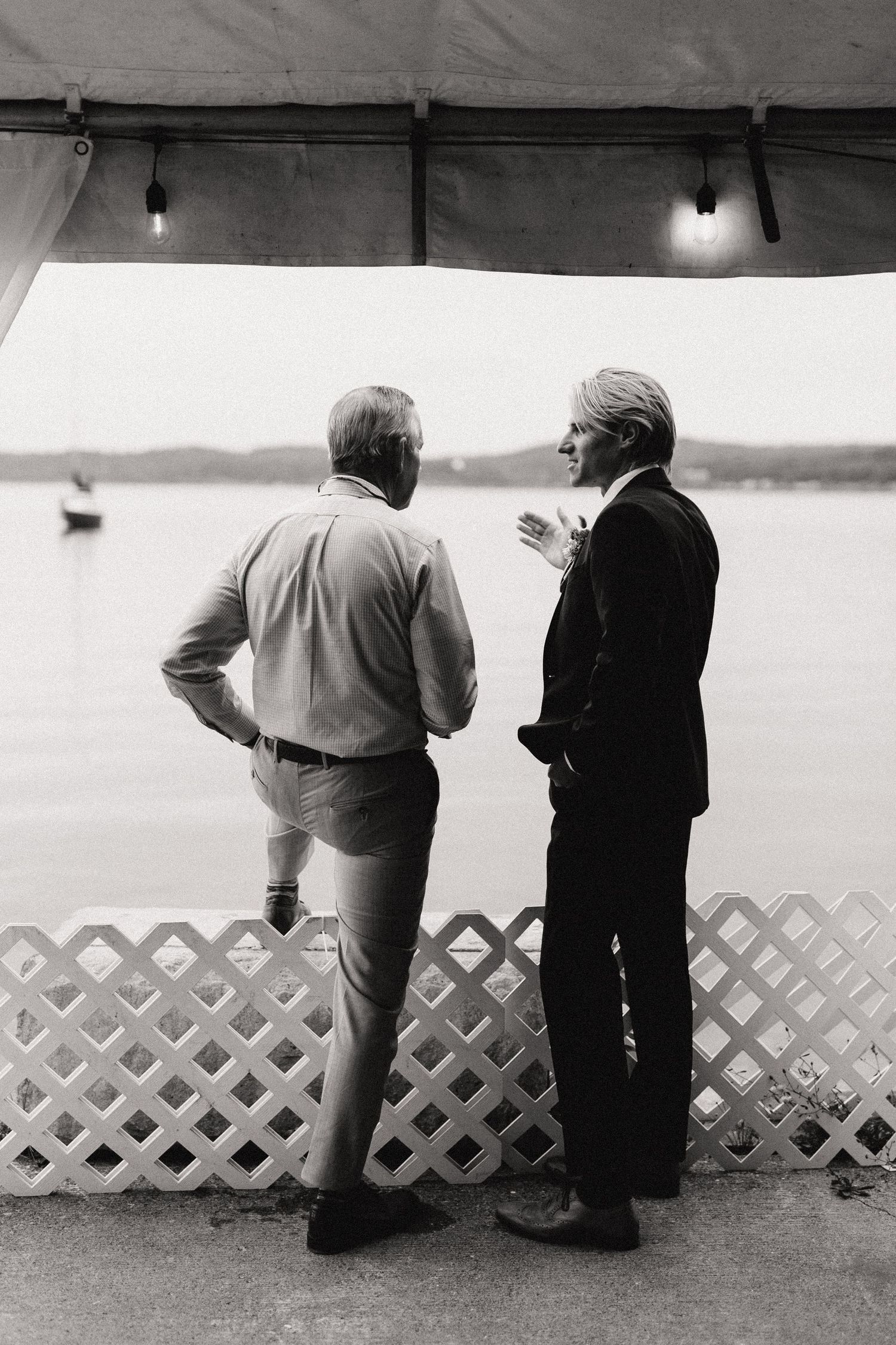 Black and white photo of two people leaning against a white lattice railing overlooking water.