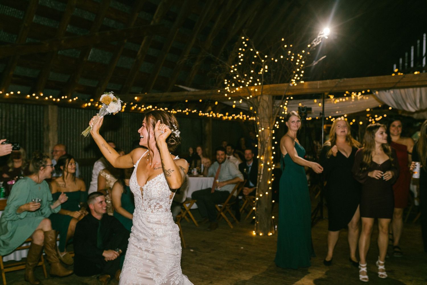 Bride in white dress dancing with guests and readying to throw the bouquet at rustic wedding reception under twinkling lights.