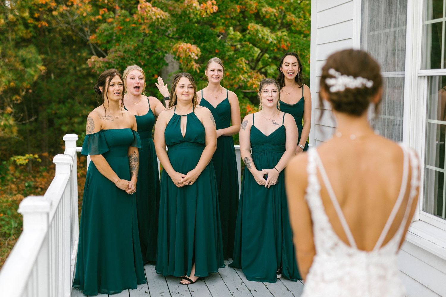 Bridesmaids in emerald green floor-length dresses stand together on a white porch during an autumn wedding.