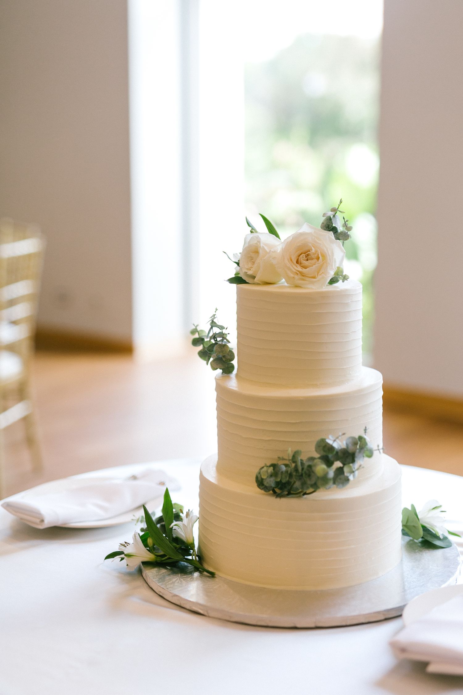 Elegant three-tier white wedding cake decorated with white roses and greenery on a reception table.