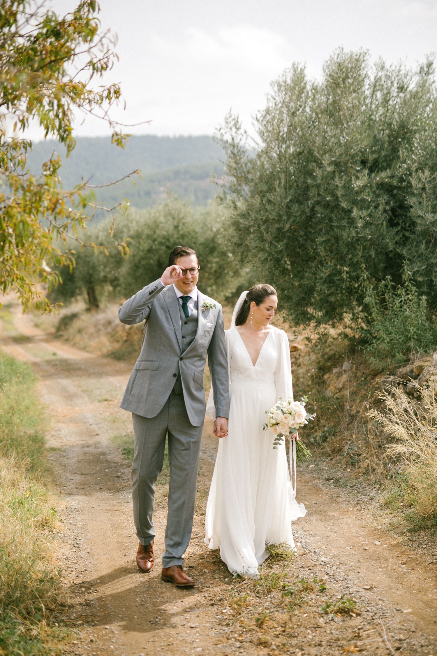 A wedding couple walks along a scenic olive grove path with hills in the background. Europe and Spain destination wedding photographer.