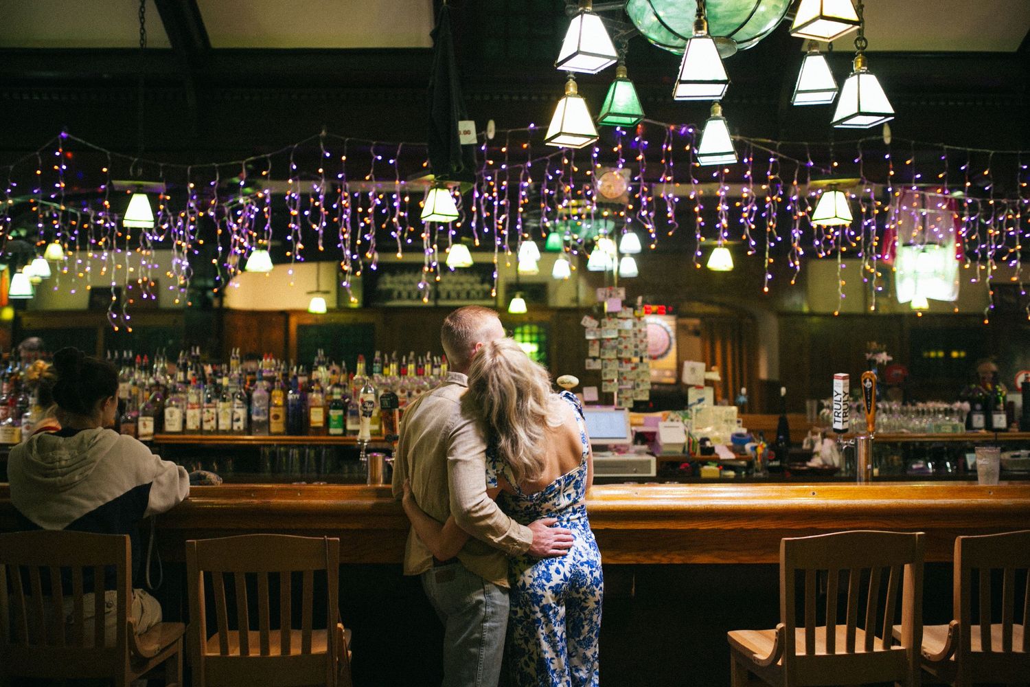 A couple shares an intimate moment at a cozy bar with twinkling lights and pendant lamps hanging overhead.