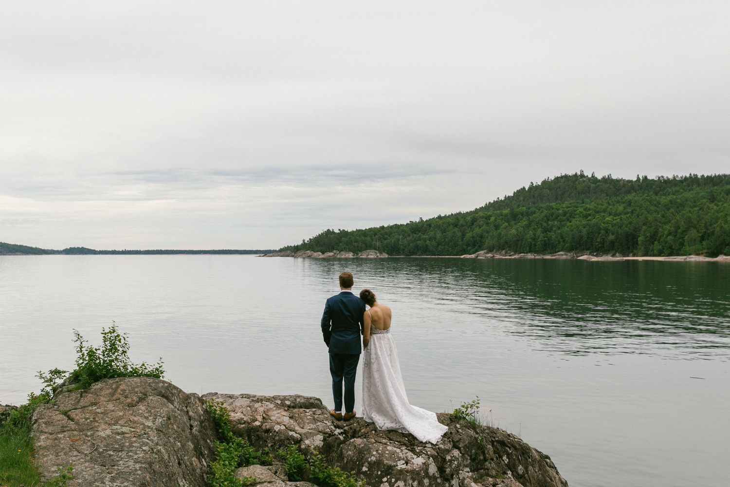 A couple in wedding attire stands on a rocky outcrop overlooking a serene lake surrounded by green forested hills.