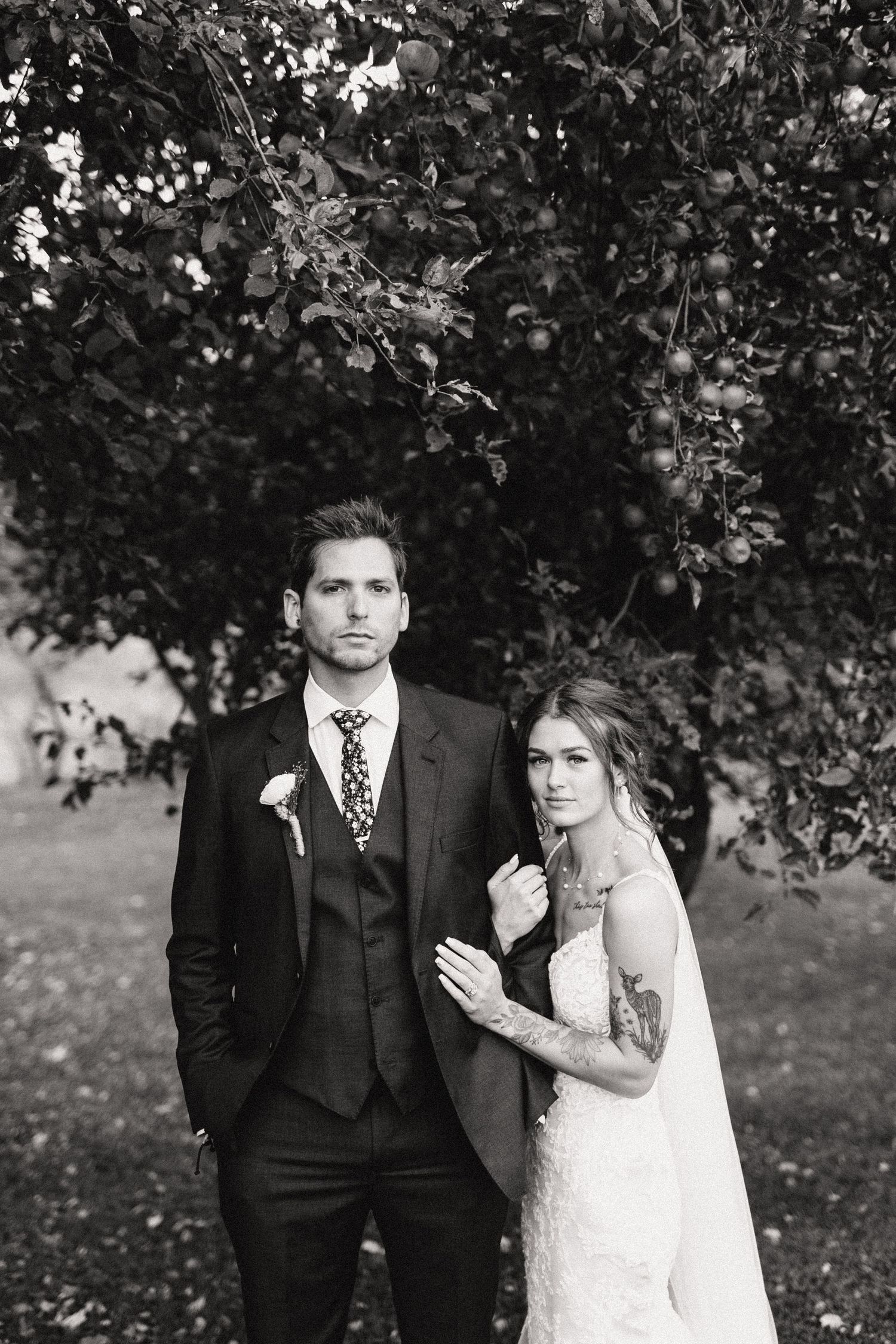A black and white wedding photo with the couple posing outdoors under lush tree branches.