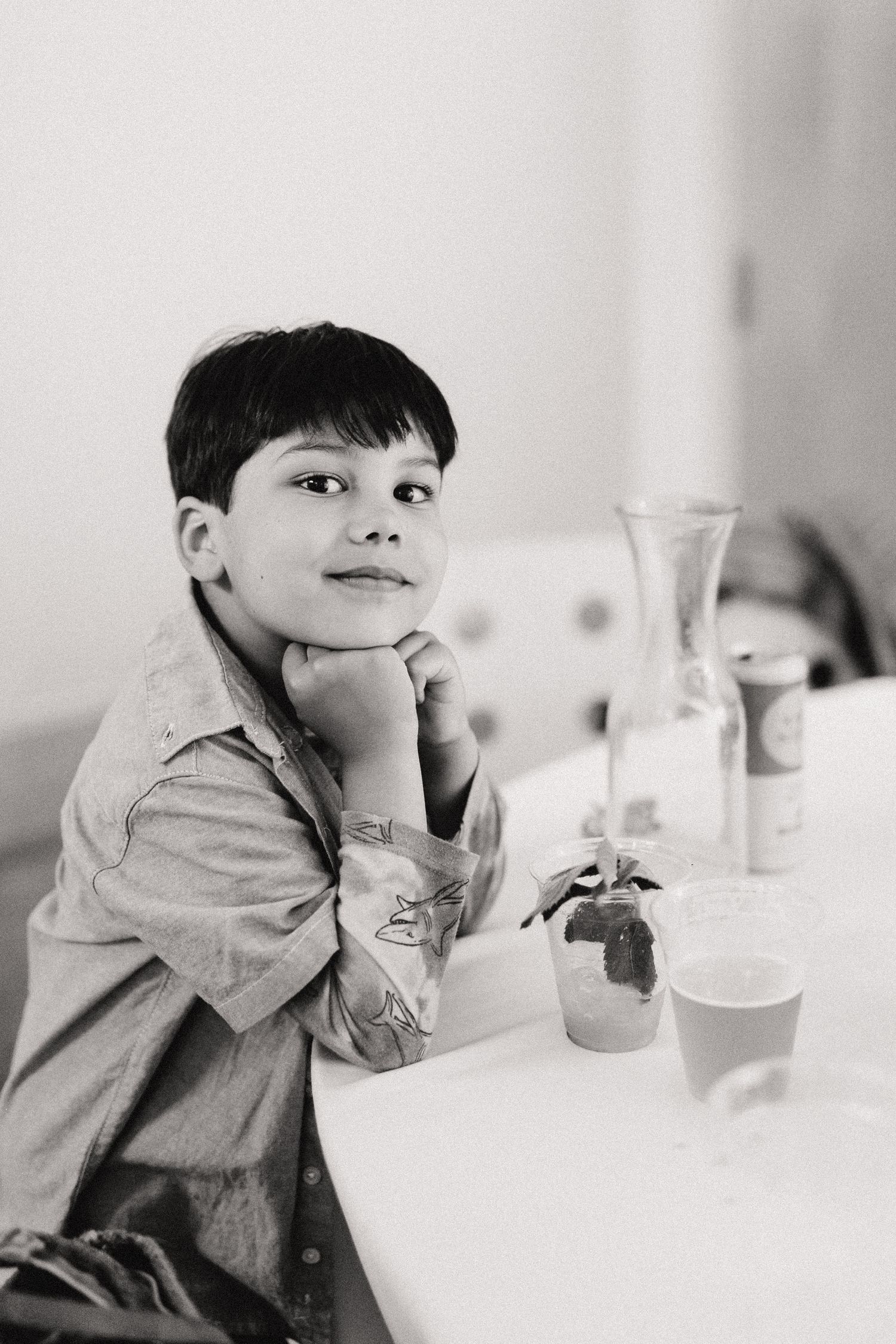 Black and white portrait of someone in a casual shirt sitting at a table with a glass vase and potted plant.