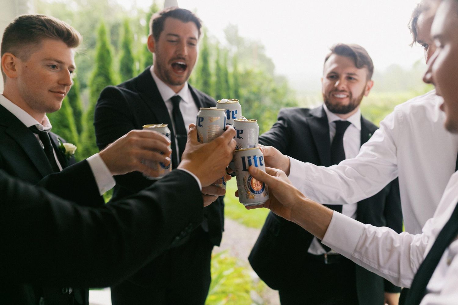 A series of groomsmen in black suits and white shirts perform a beer toast outdoors.