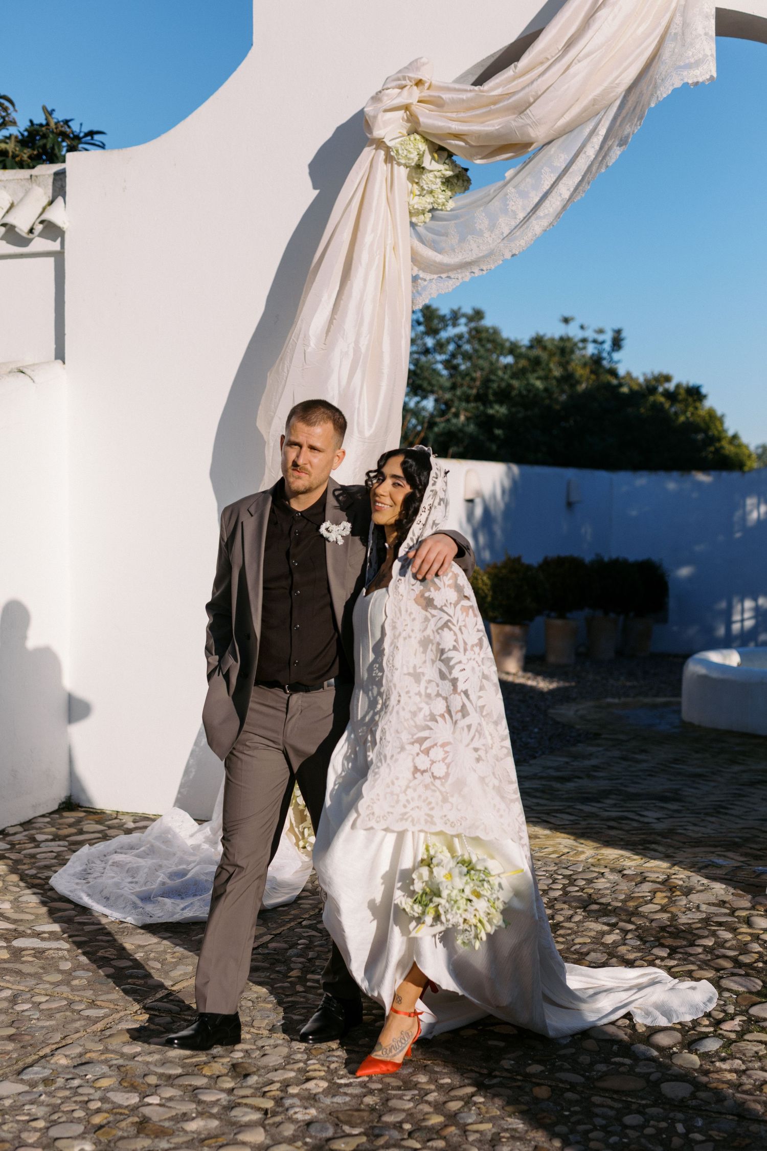 Bride in lace mantilla veil with orange heels leans on groom beside white arch at sunny venue.