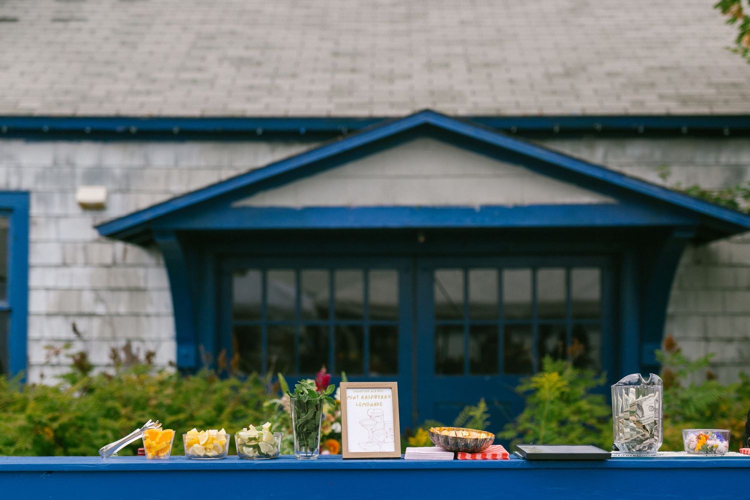 A rustic outdoor buffet table with food and drinks set up in front of a blue-trimmed house with shingle siding.