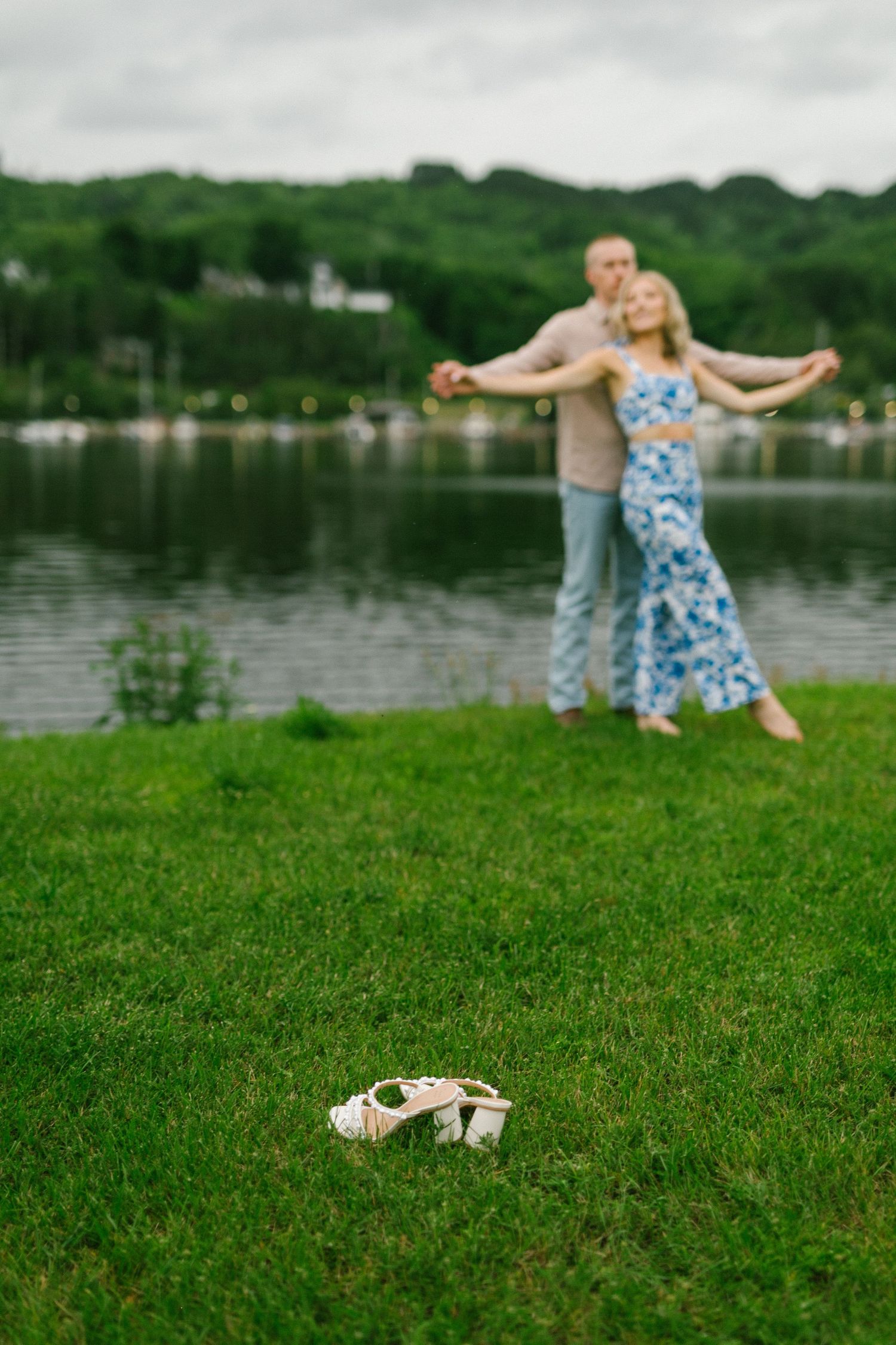A couple dances together on grassy lakeside lawn with sandals in foreground on summer afternoon.