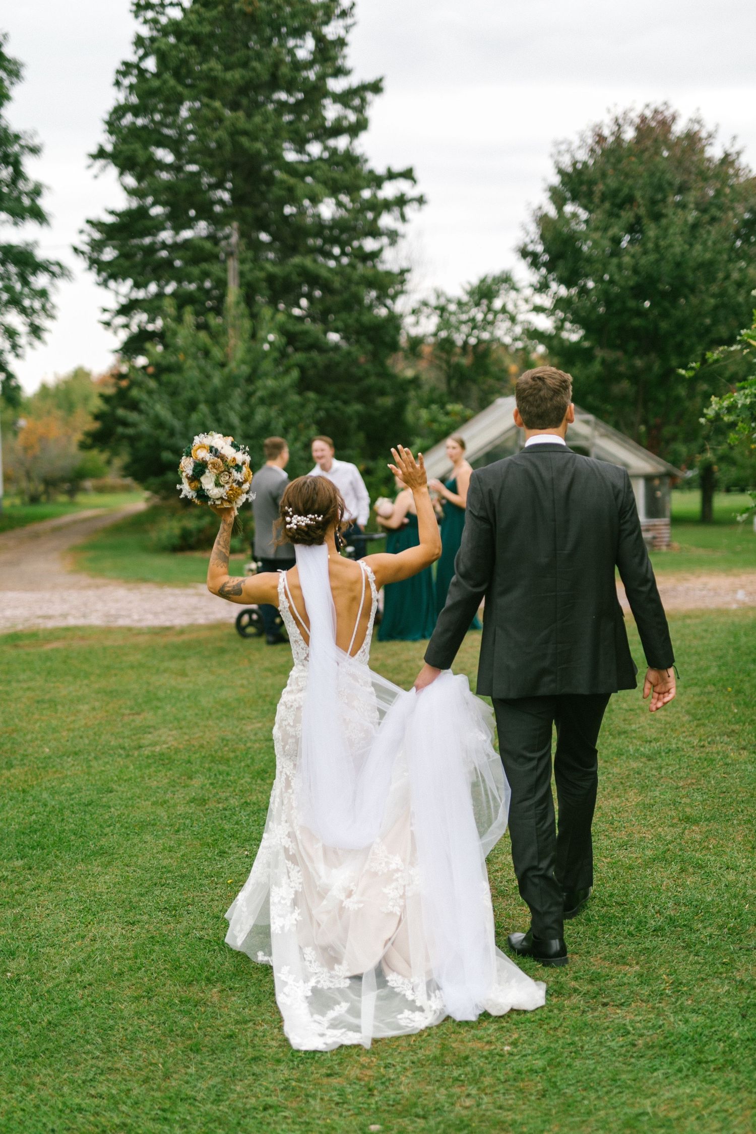 A celebratory outdoor wedding scene with the couple walking on a grassy path surrounded by green trees.
