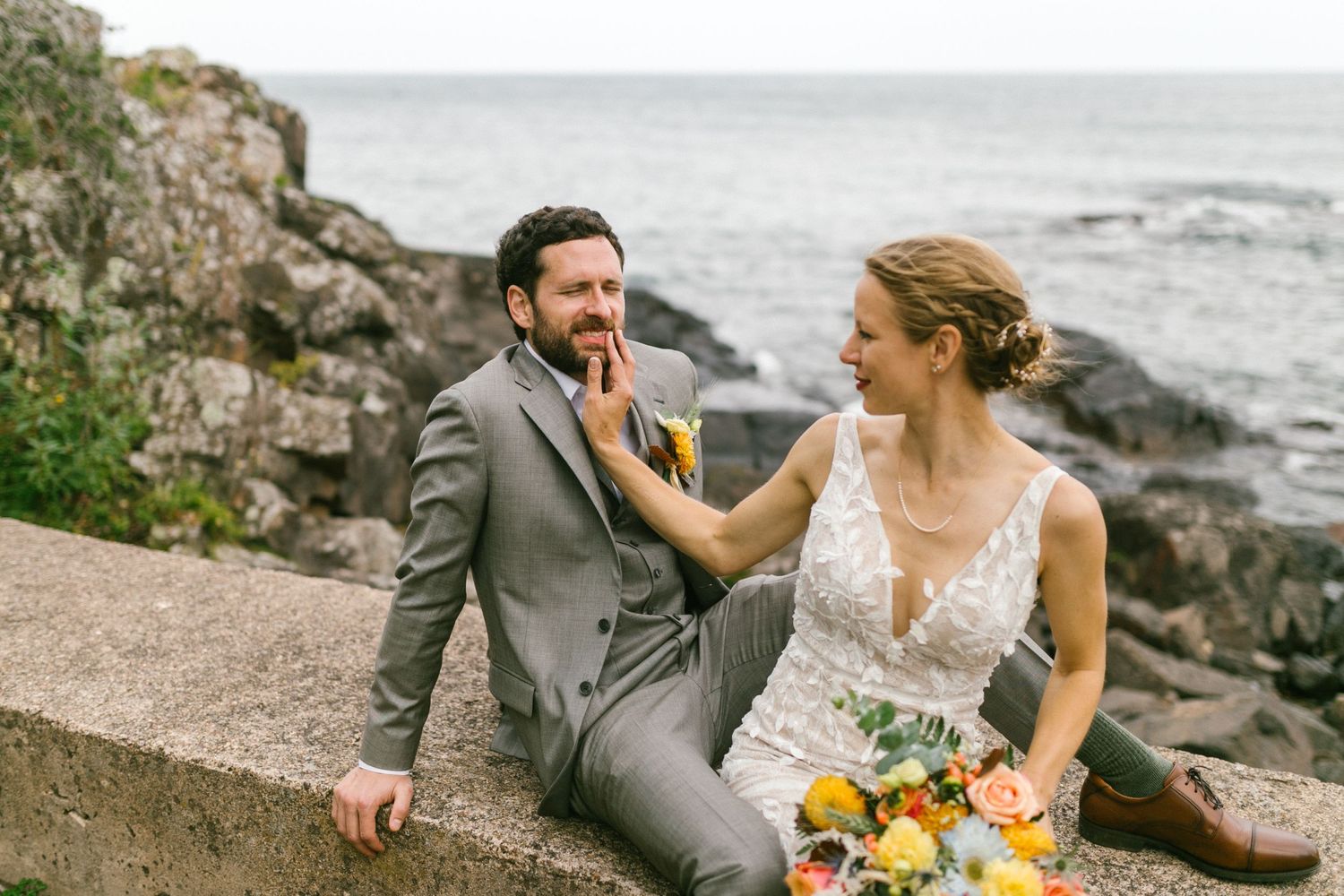 A couple in wedding attire share a playful moment on rocky coastal cliffs with a bouquet of yellow flowers.
