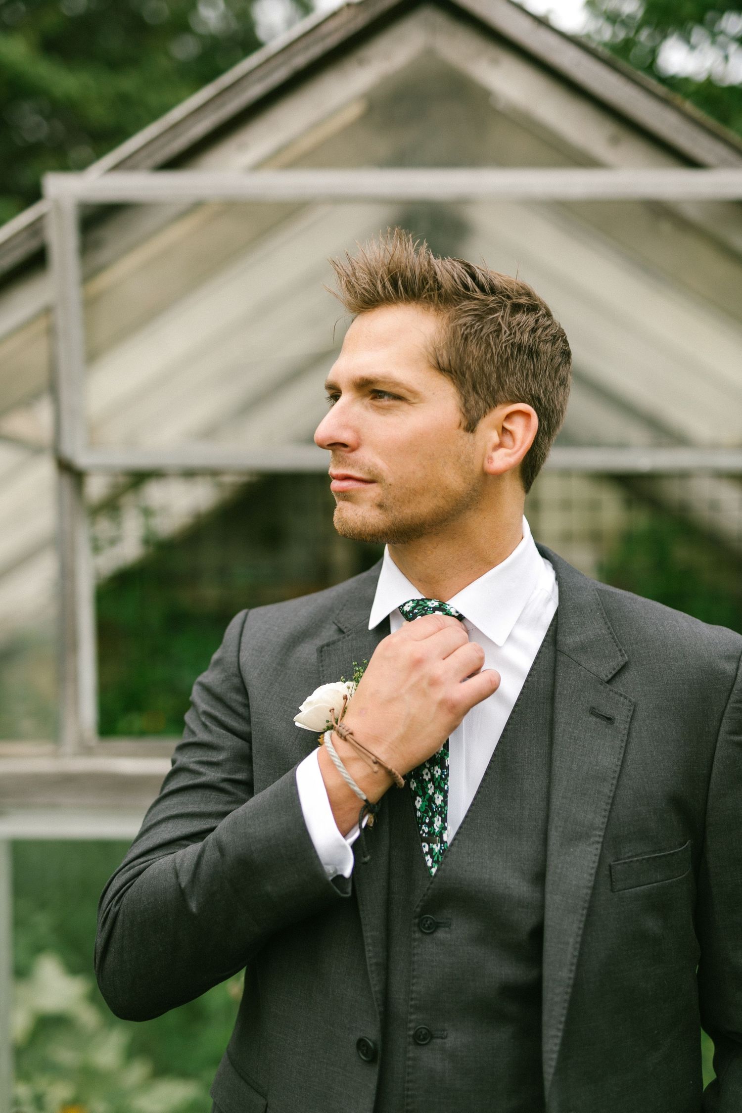 Groom adjusts his polka dot tie while wearing a dark gray suit in front of a greenhouse structure.