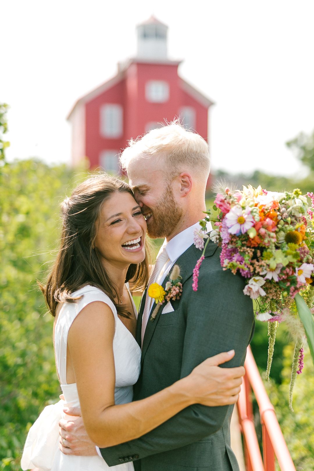 Happy couple embraces in front of red lighthouse with vibrant summer flower bouquet.