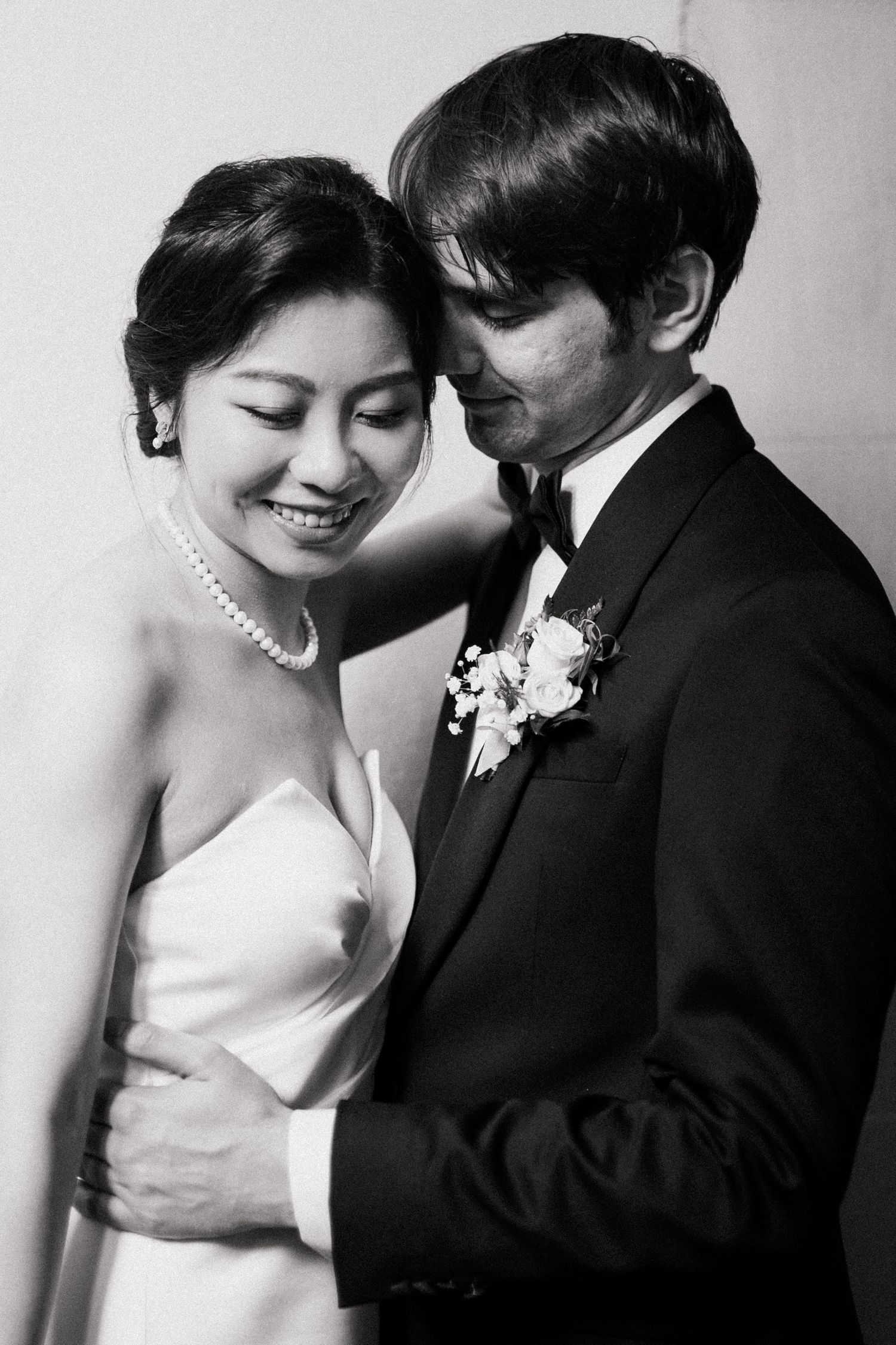Black and white portrait of a smiling bride in pearls embracing her groom in a tuxedo.