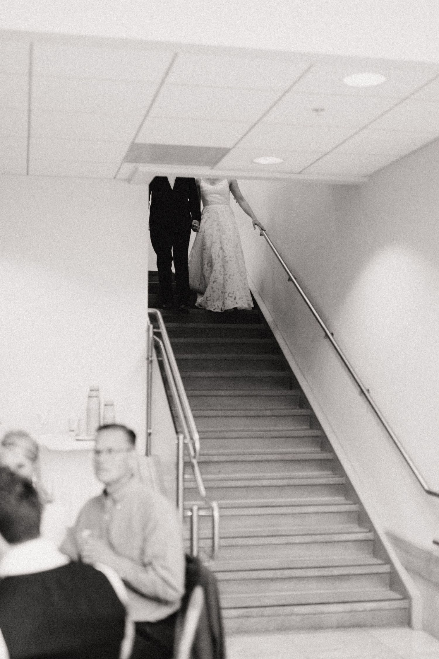 A black and white photo showing a bride and groom descending a staircase with metal handrails and white walls.