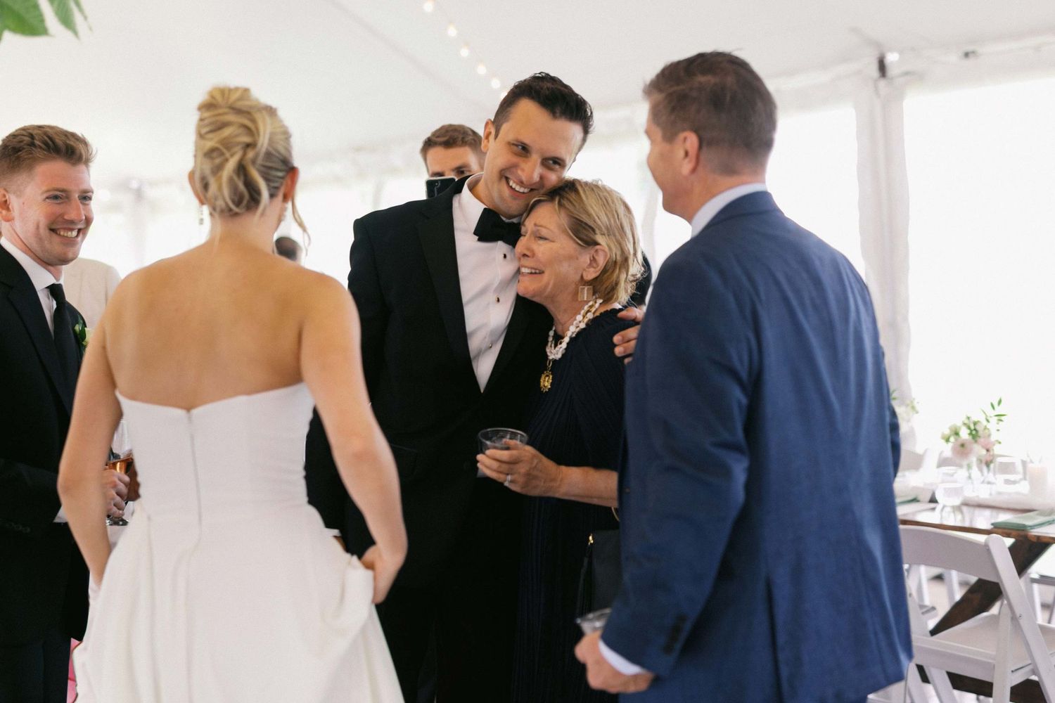A group of formally dressed wedding guests and attendees gather under a white tent during a celebration.