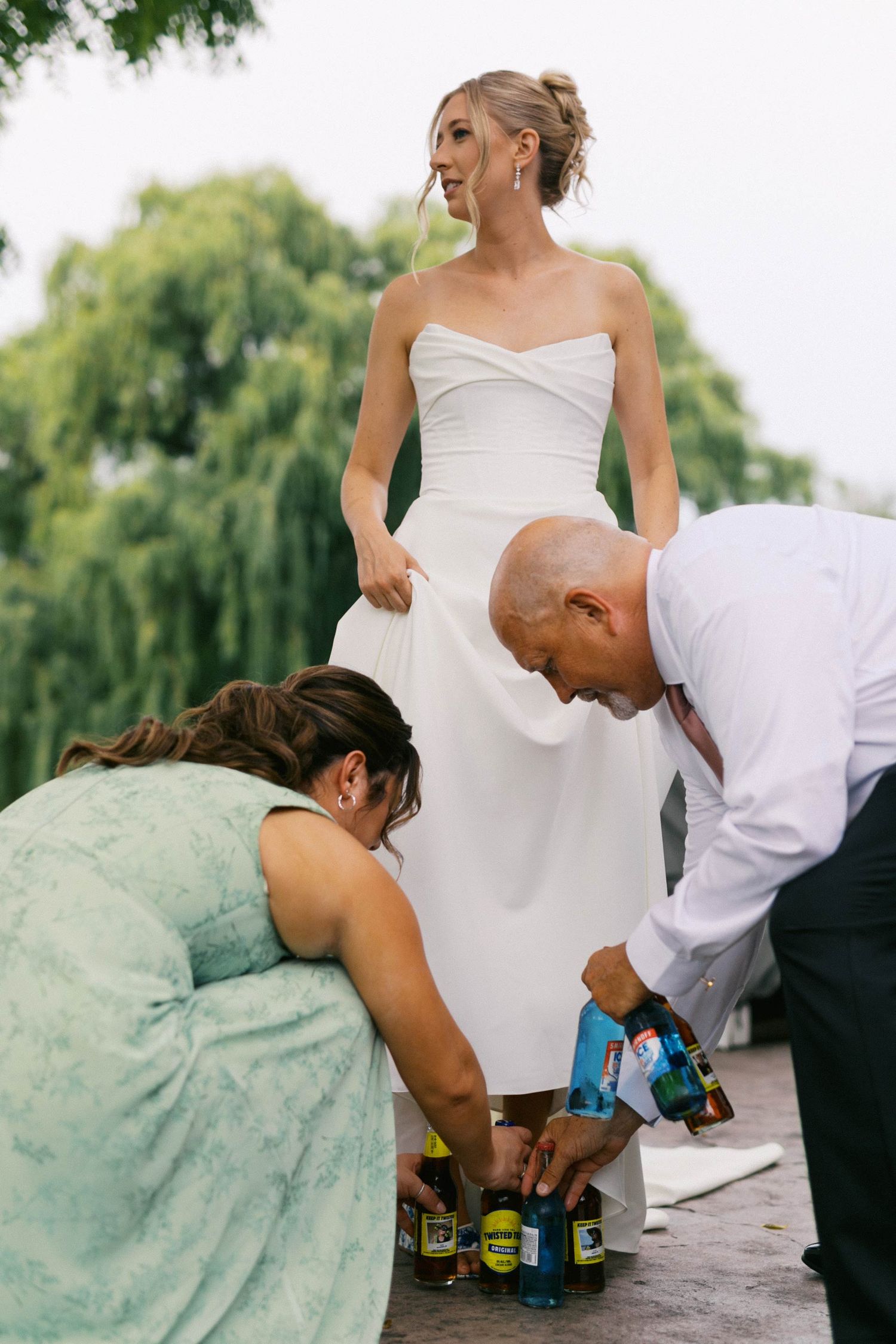 A bride in a white dress stands while someone in mint green and another in white help with a surprise for the groomsmen.