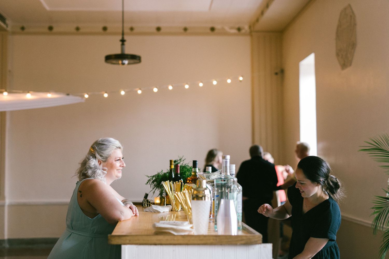 People gather at a modern bar counter with string lights overhead and natural light streaming through windows.
