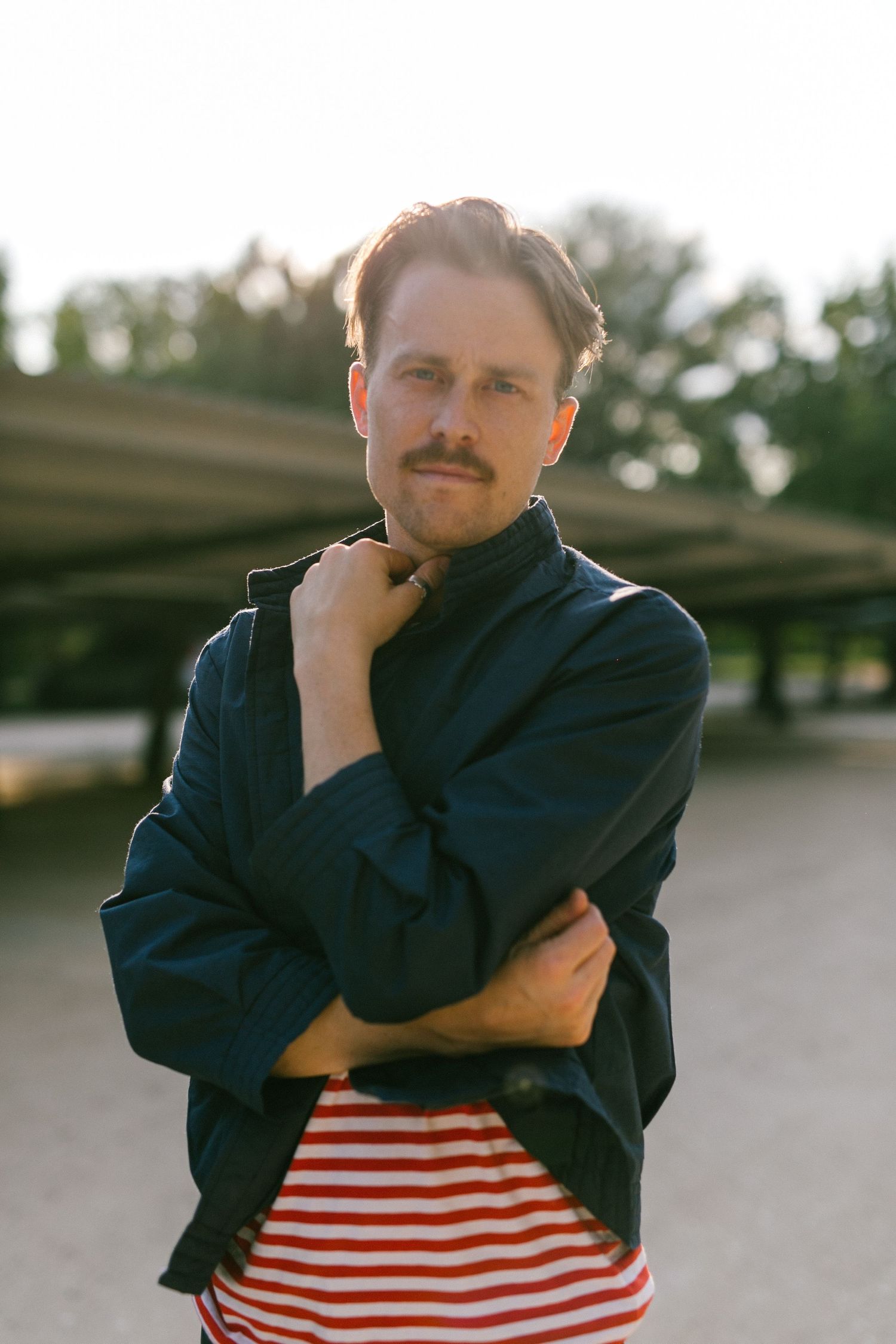 Person in navy jacket and red striped shirt poses with crossed arms at dusk.