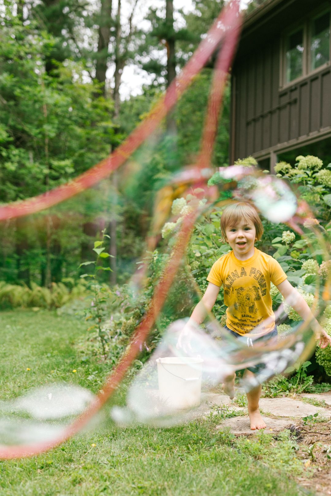Excited young boy in yellow shirt playing with giant soap bubbles in green backyard.