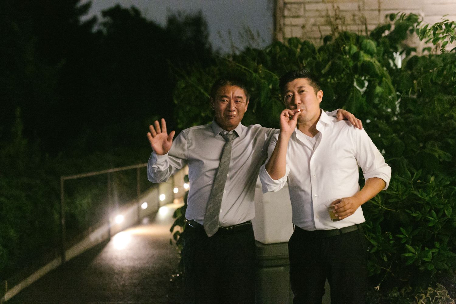 Two Asian men smiling and posing outdoors at night, one waving and one on phone, near lit pathway.
