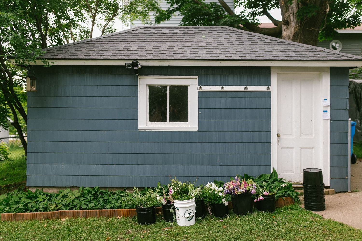 A small blue-gray garden shed with white trim and potted flowers along the foundation.