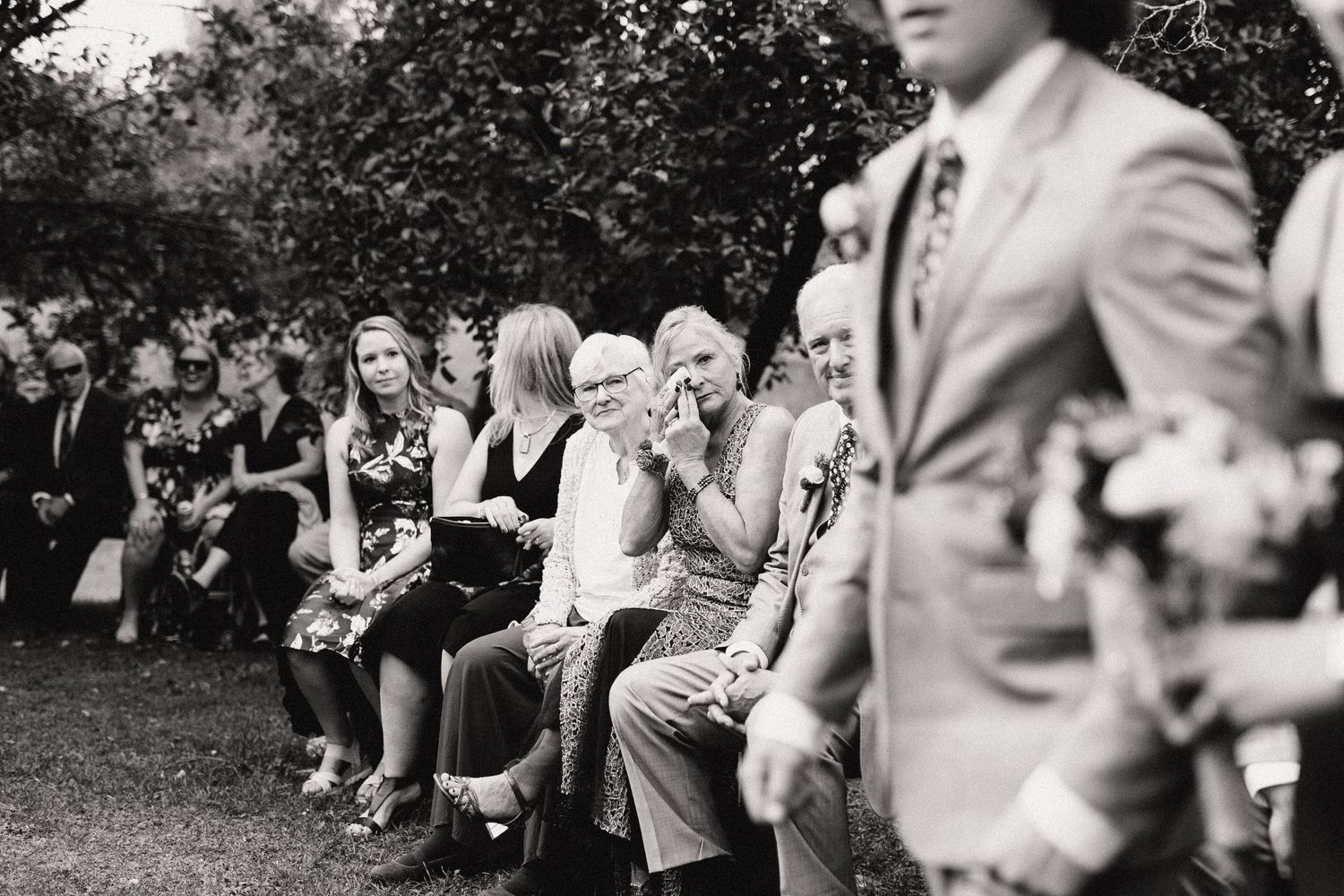 Black and white photo of wedding guests seated in rows outdoors watching the ceremony. Mother of groom dabbing tears during ceremony.