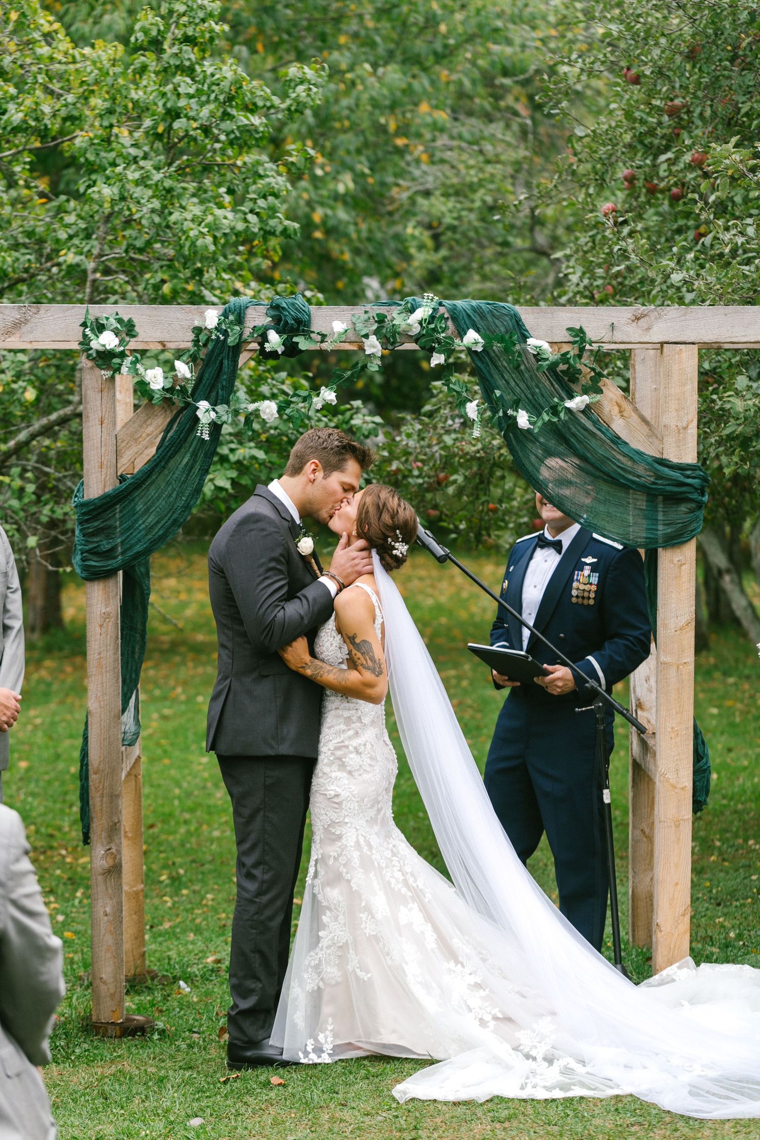 A wedding ceremony under a wooden arch decorated with emerald green fabric and white flowers in a garden setting.