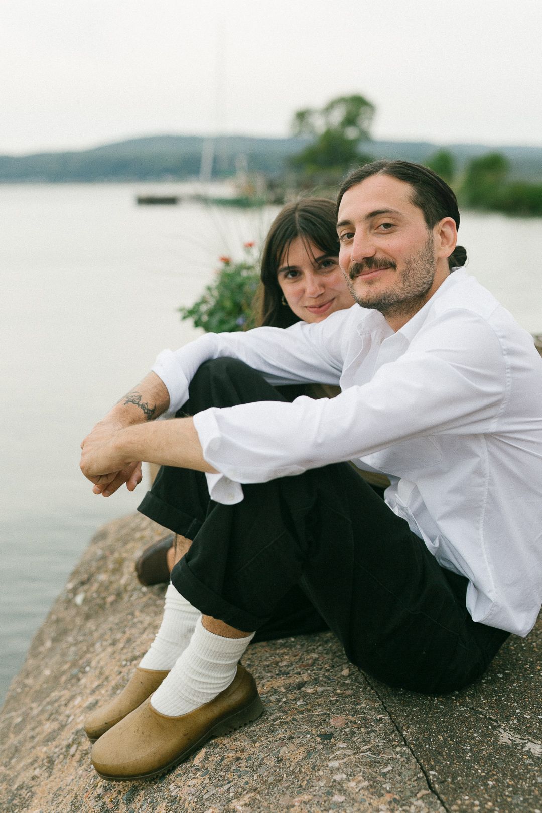 Couple sitting together on rocky coastline overlooking peaceful water.