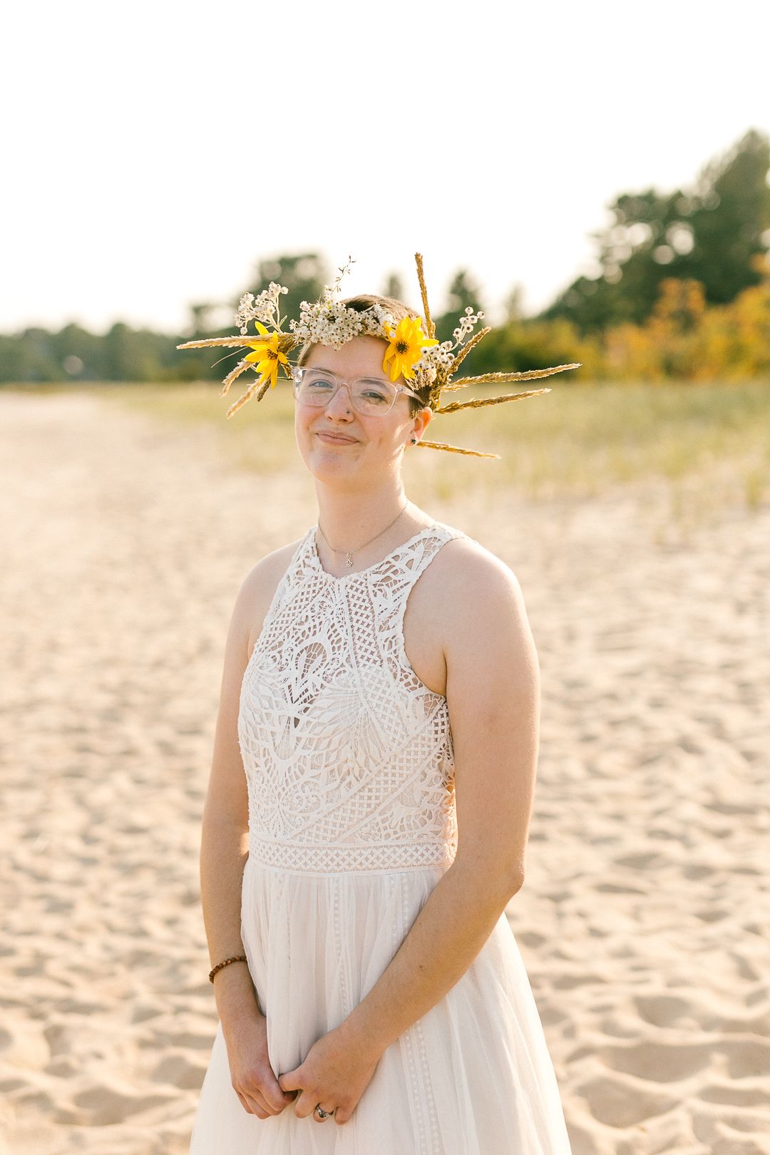 A person in a white lace dress poses on a sandy beach with sunflowers at sunset.