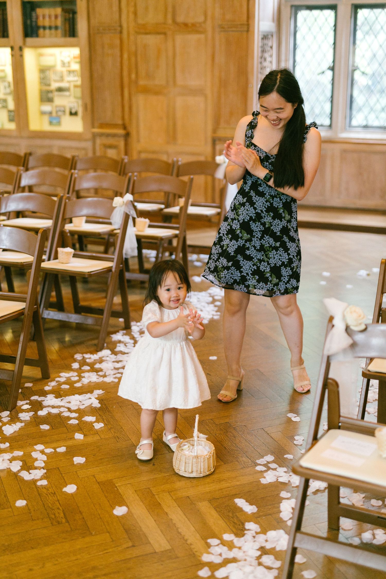 Adorable flower girl in white dress scatters rose petals down wedding aisle with smiling woman in floral dress.
