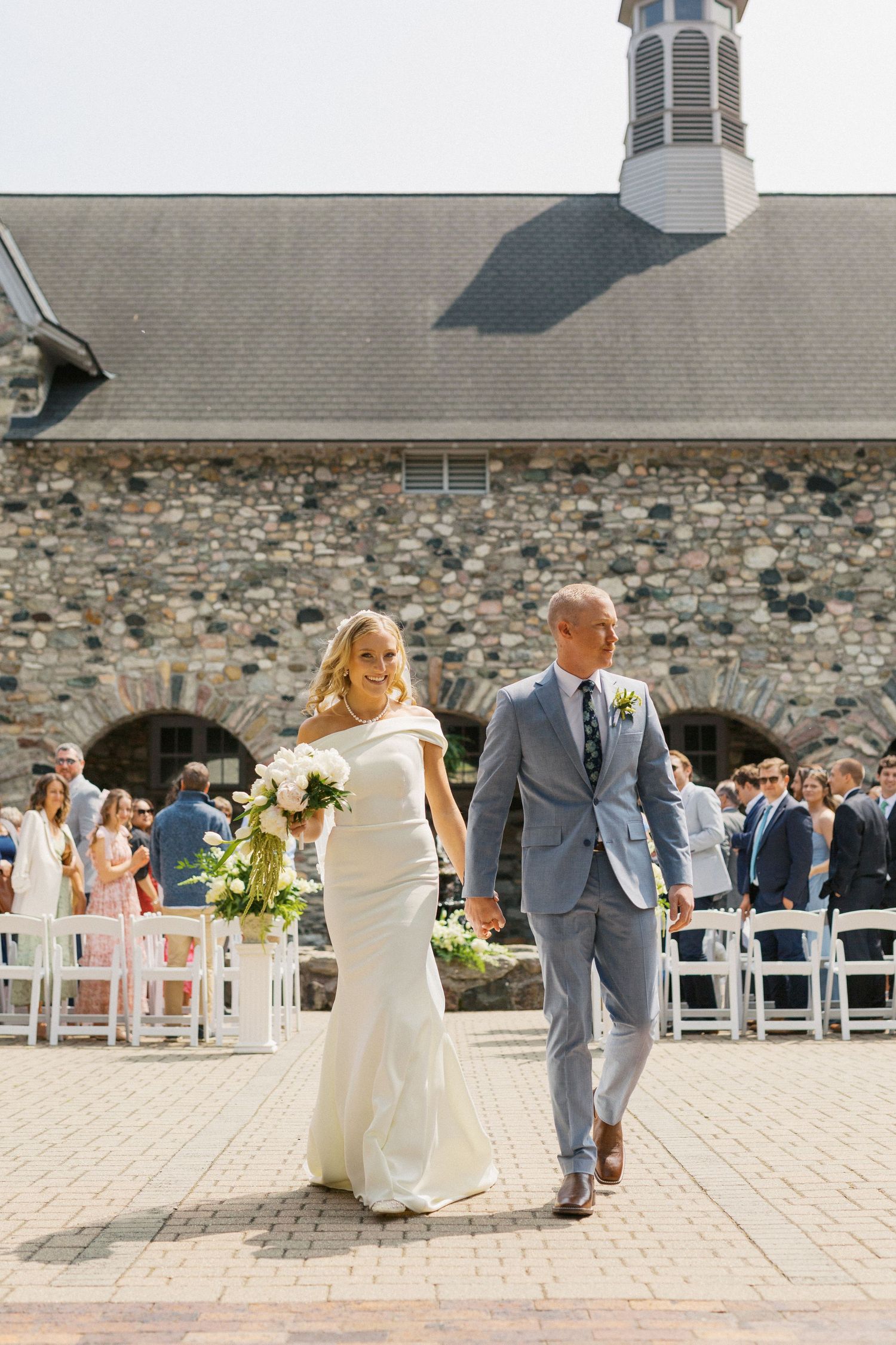 A wedding couple walks together after their ceremony outside a historic stone church with white chairs set up on the patio.