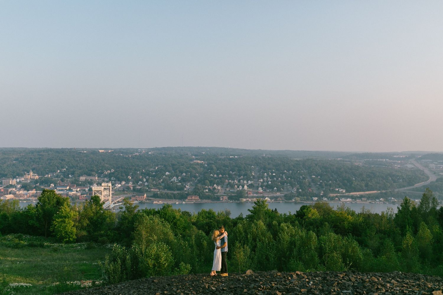 A distant view of a couple embracing on a hilltop overlooking a scenic landscape at dusk.