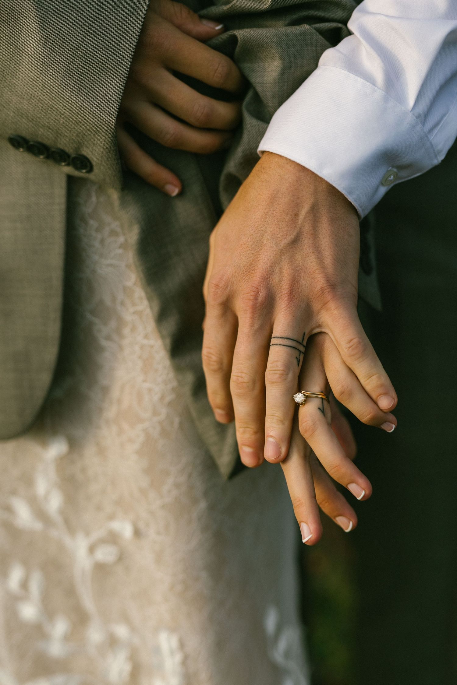 Close-up view of newlyweds' hands displaying their wedding rings against formal attire and white lace.