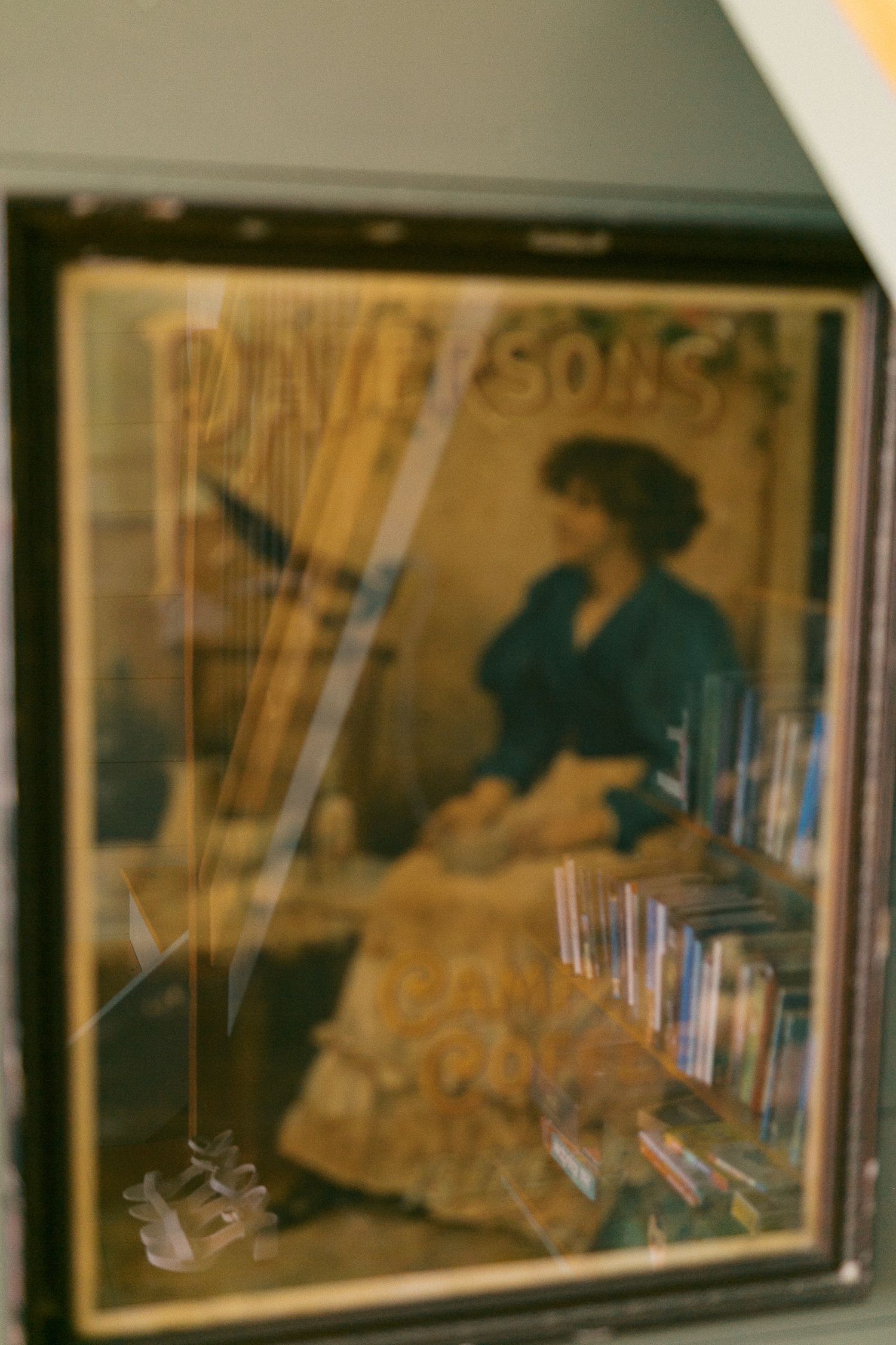Blurry vintage poster of a woman playing harp displayed in a glass case with books and wedding shoes reflected in the glass.