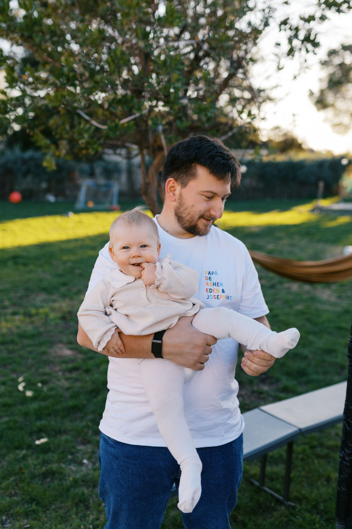 Smiling father in white t-shirt holding his baby outdoors in a green backyard on a sunny day.
