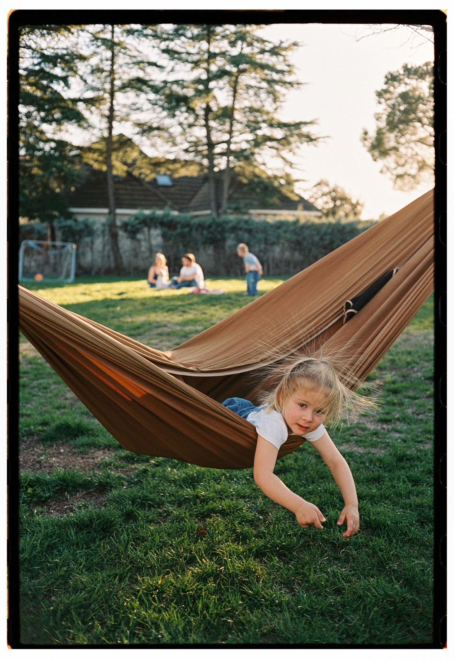 Young girl lying in a brown hammock in a sunny backyard, with children playing on grass in the background.