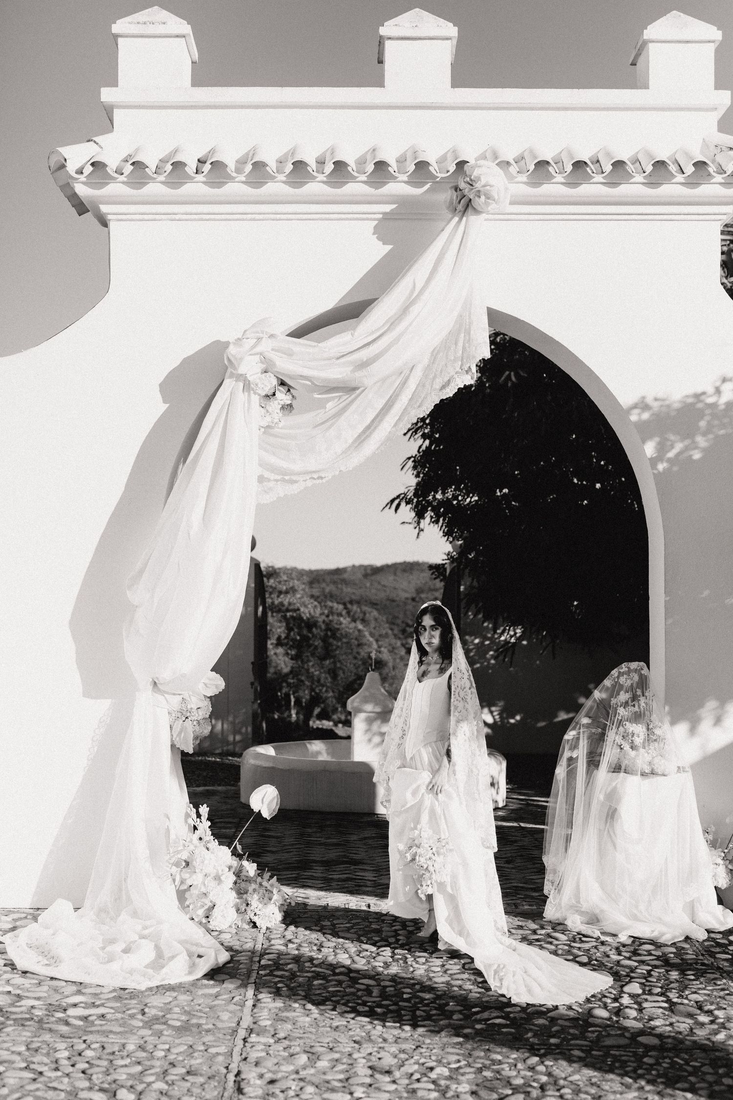 Black and white photo of bride in cathedral veil standing under draped white arch at Mediterranean venue.