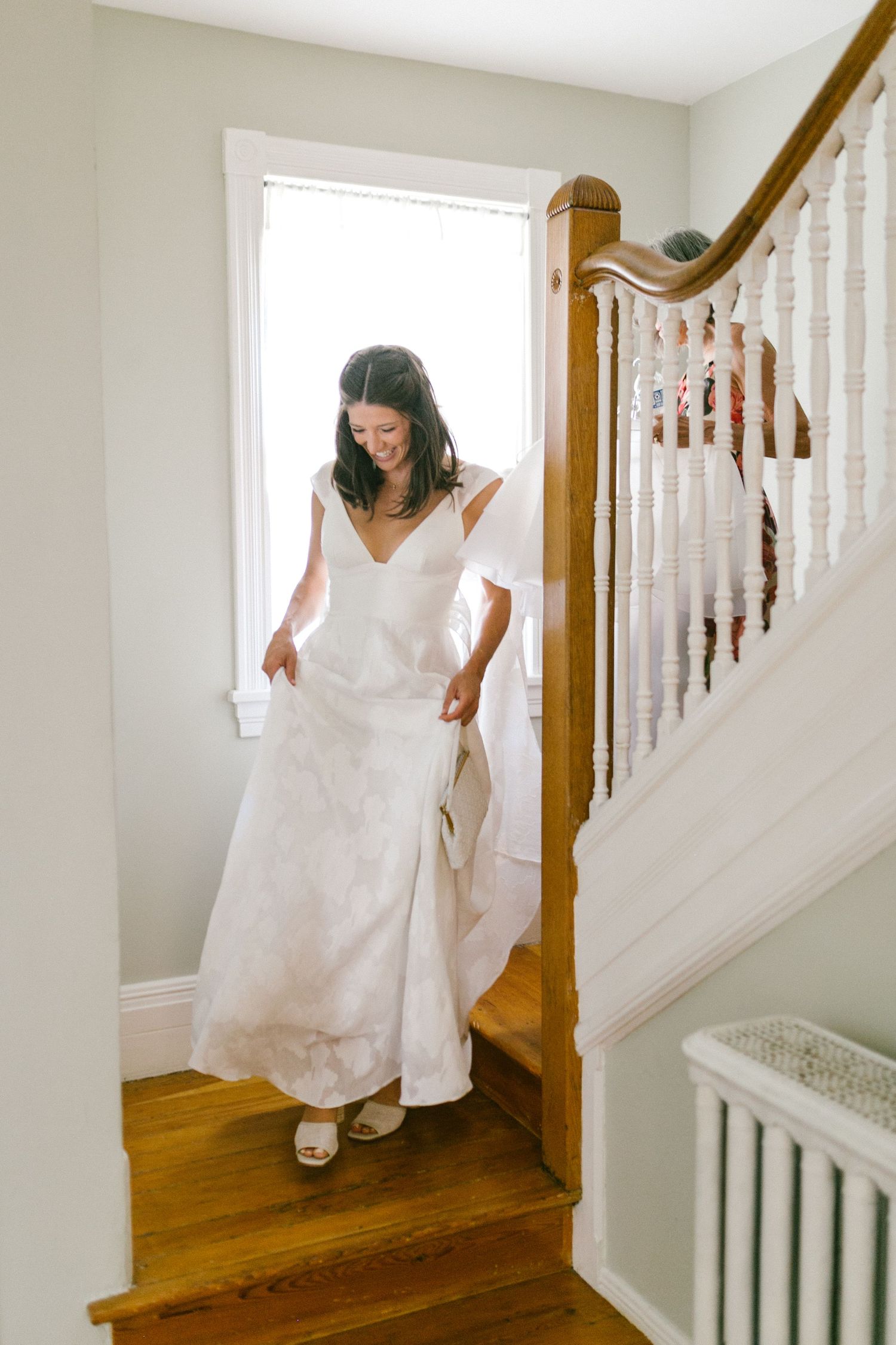 A bride in a white wedding dress descends a wooden staircase with white railings in a bright interior space.