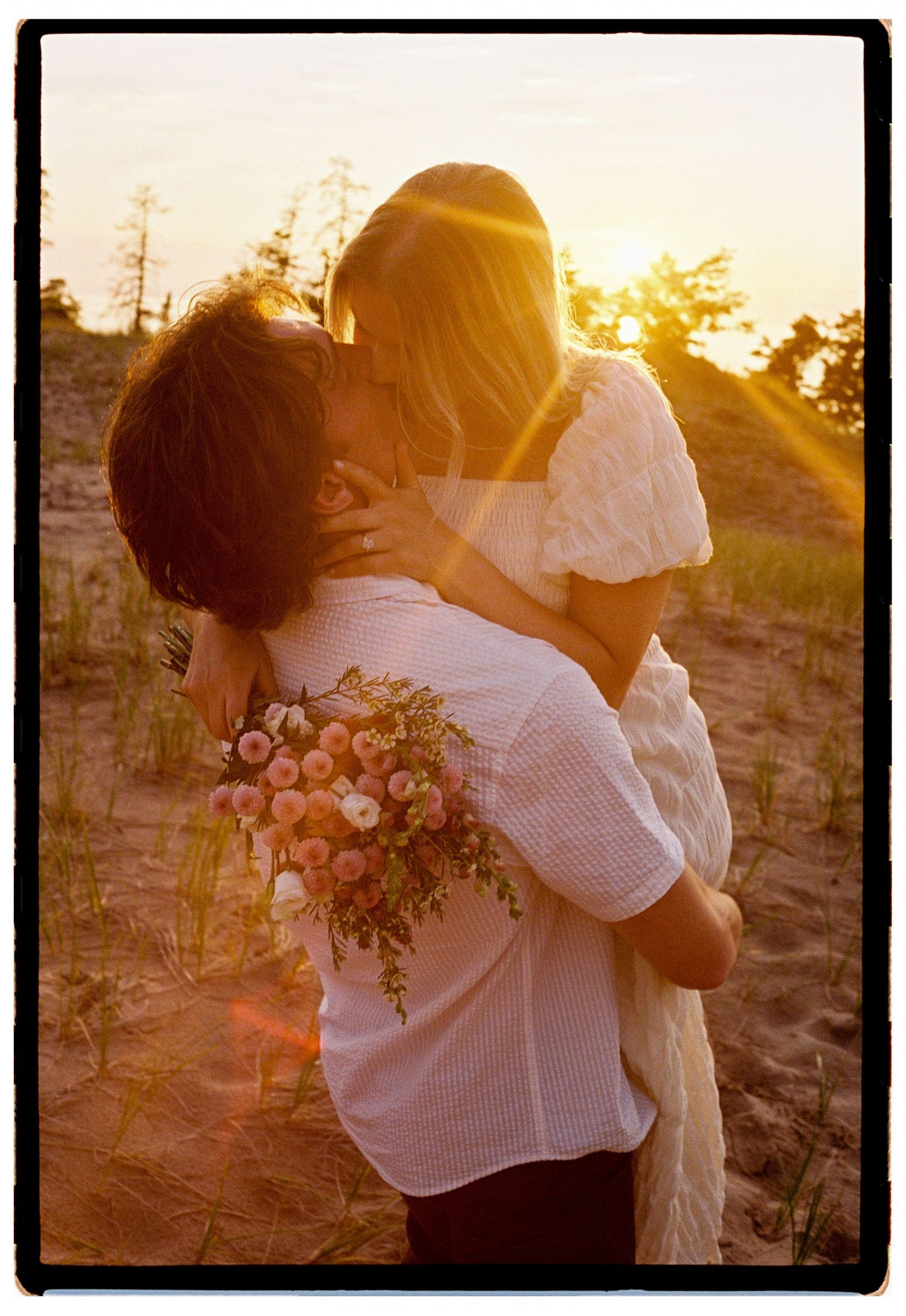 Couple sharing a romantic kiss at sunset, man holding woman and a bouquet of pink flowers on a sandy beach.