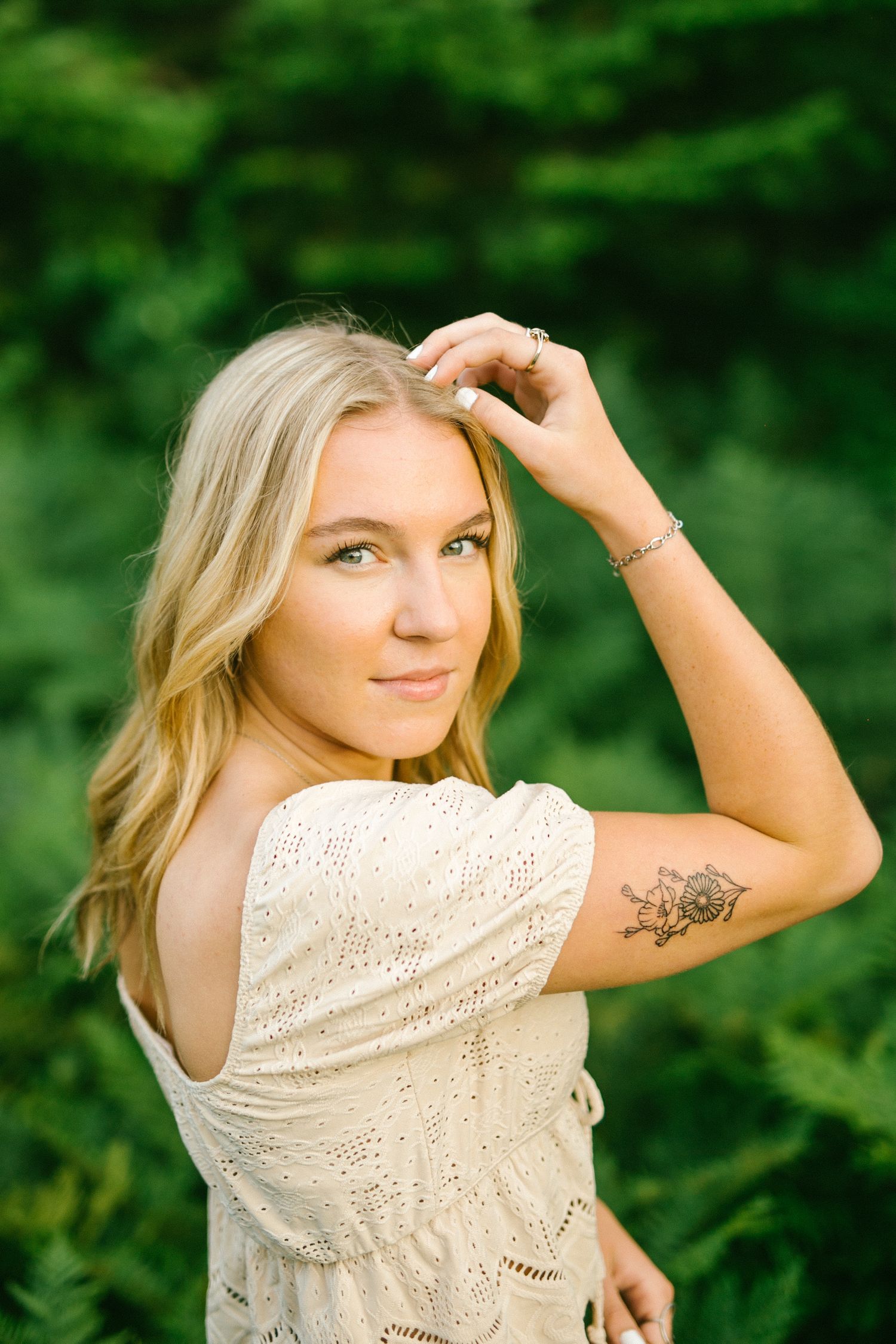 Blonde young woman with floral arm tattoo in off-shoulder cream top poses gracefully among green foliage outdoors.