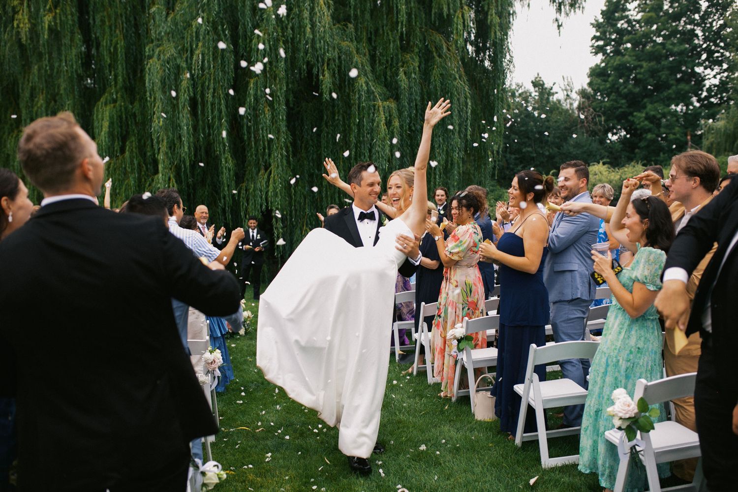 Joyful bride and groom exit wedding ceremony under flower petal toss beside weeping willow tree.
