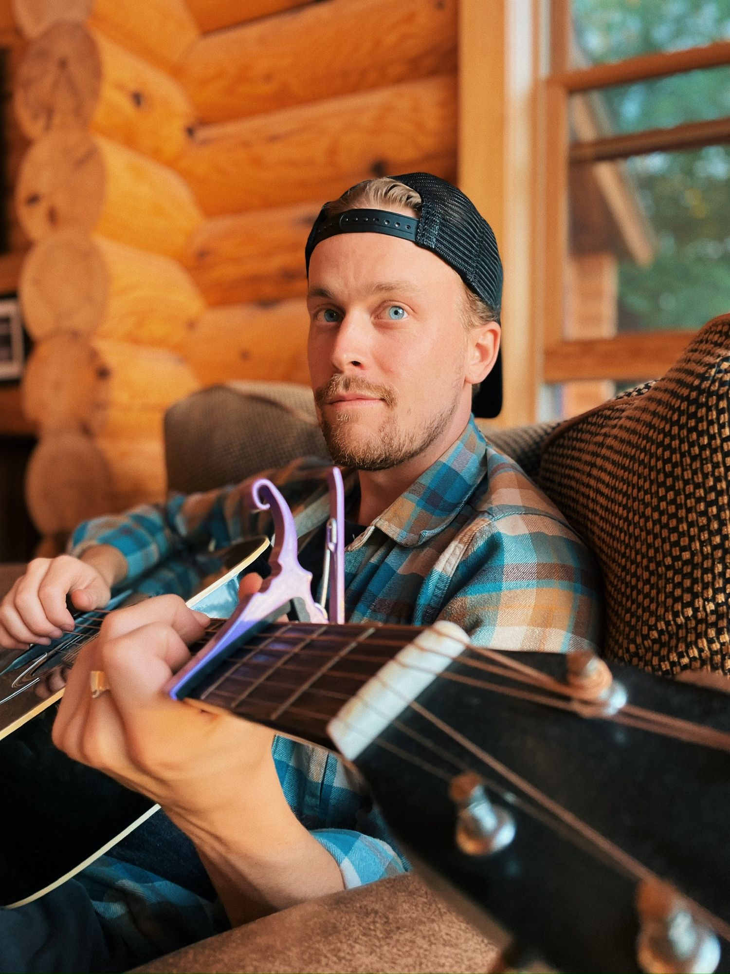 Bearded man with blue eyes playing acoustic guitar with capo in a cozy log cabin interior.
