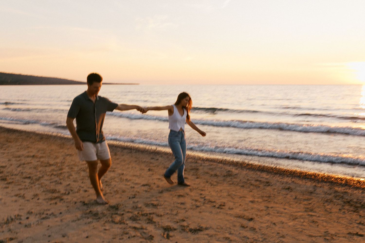 Happy couple holding hands and walking along a sandy beach at golden hour sunset.