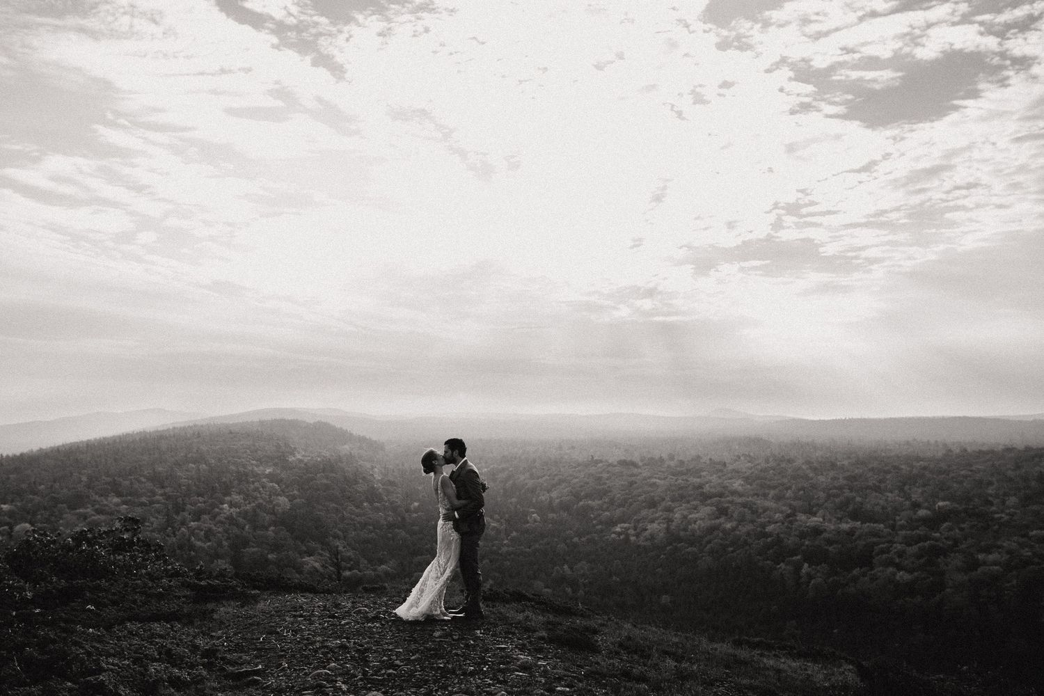 A couple in wedding attire stands silhouetted on a hilltop against a dramatic cloudy sky overlooking a vast landscape.