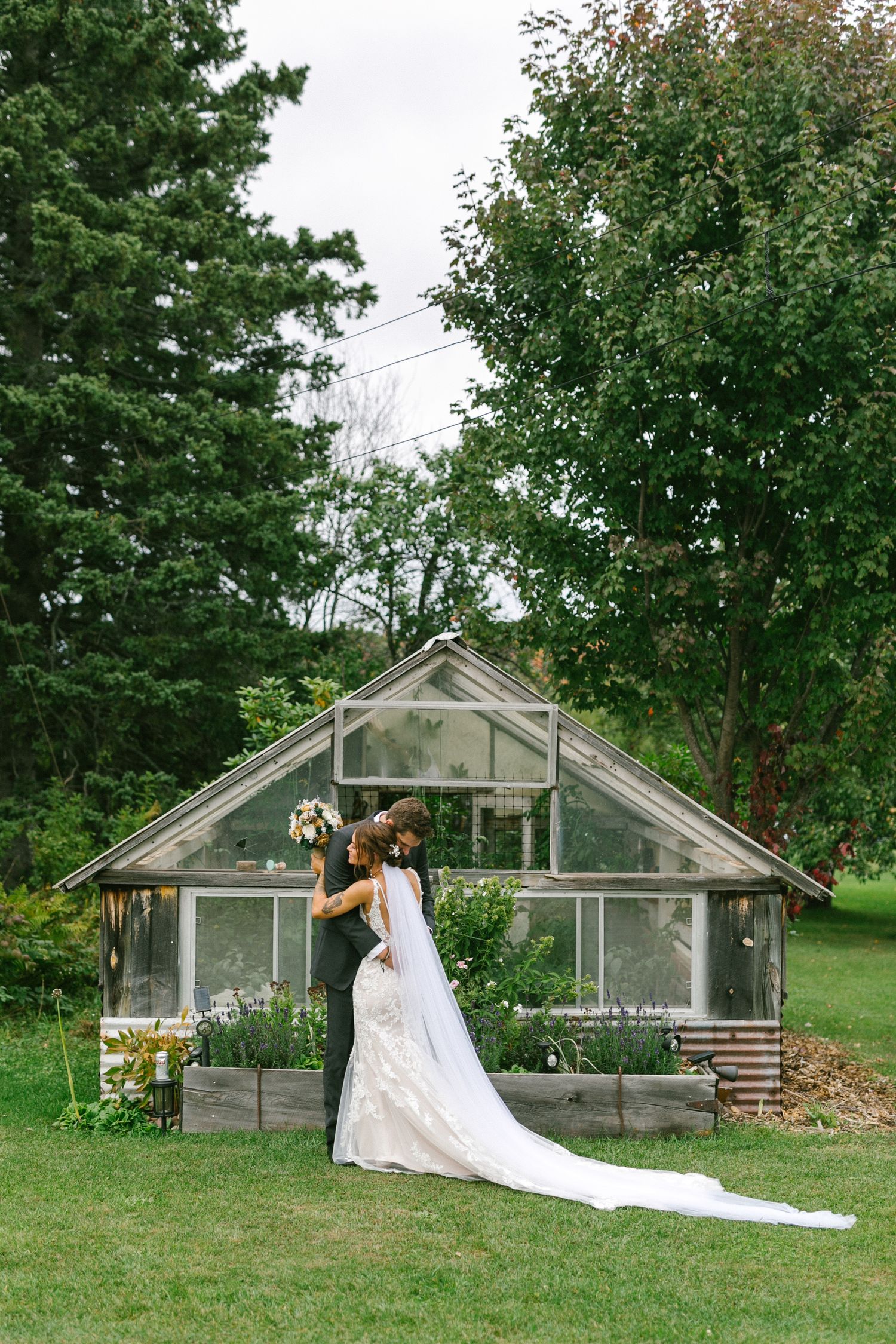 A wedding dress train trails elegantly behind someone standing near a rustic greenhouse structure in a lush garden setting.