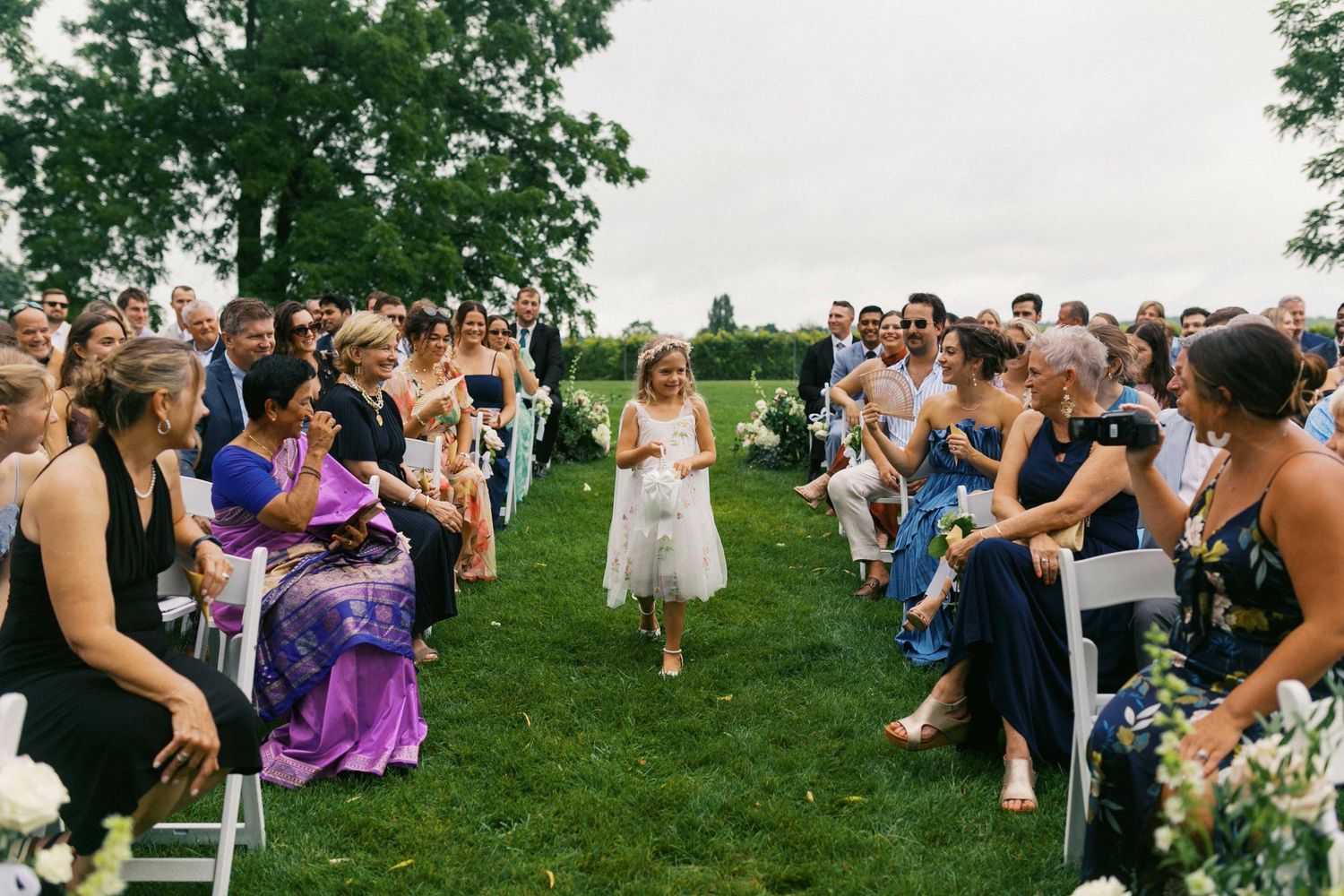 Wedding guests line both sides of a grassy aisle as a flower girl walks down during an outdoor ceremony.