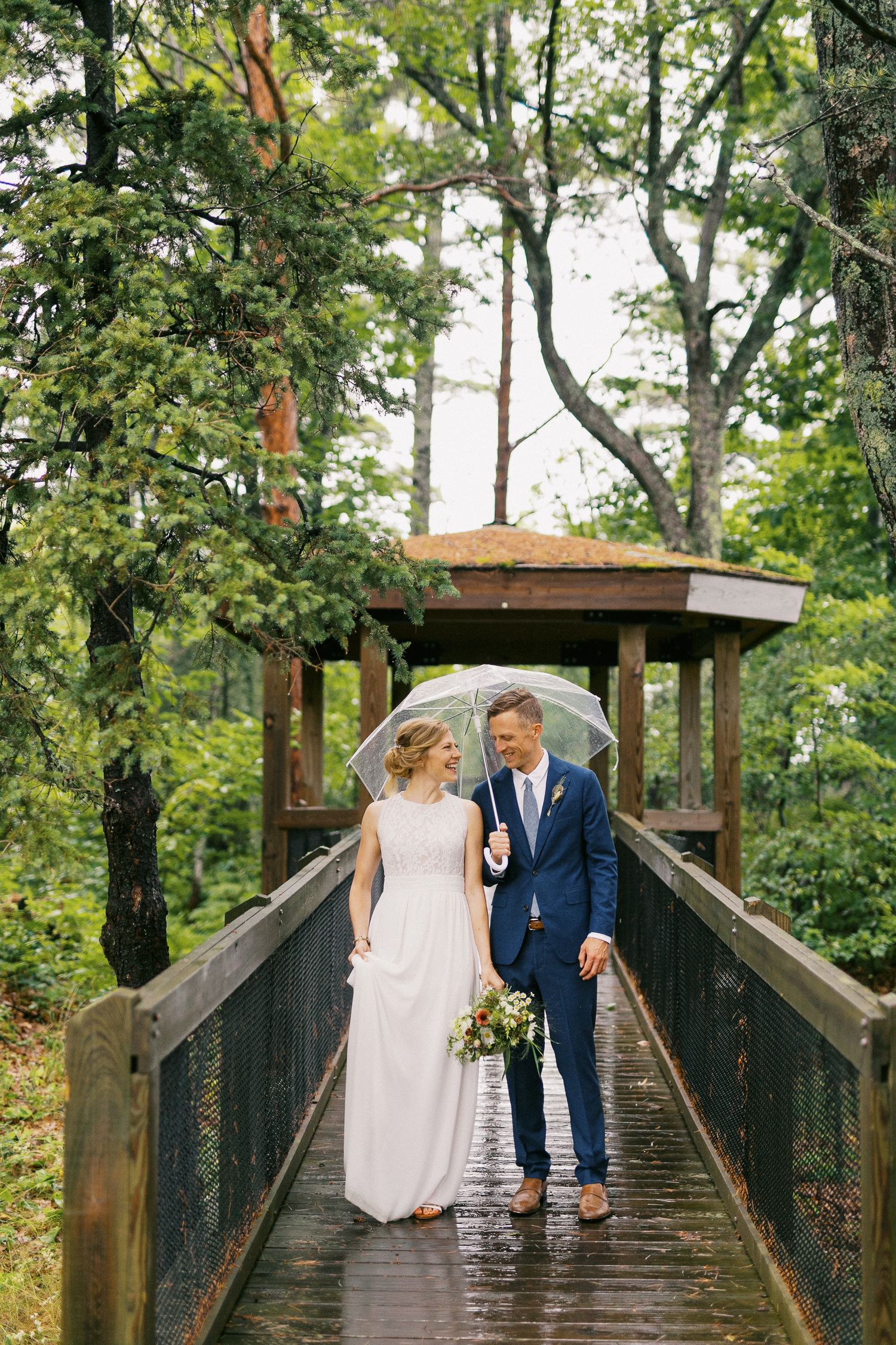 Bride and groom walking on wooden bridge in rain sharing clear umbrella in lush forest setting.