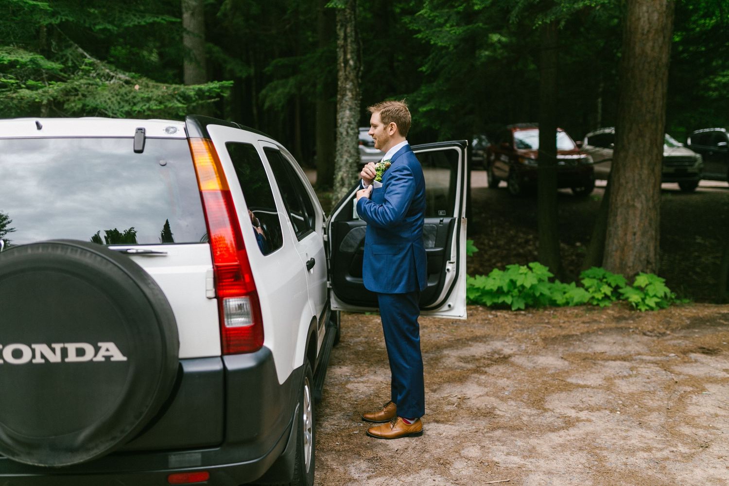 Someone in a blue jacket stands by a white Honda SUV in a wooded area on a rainy day.